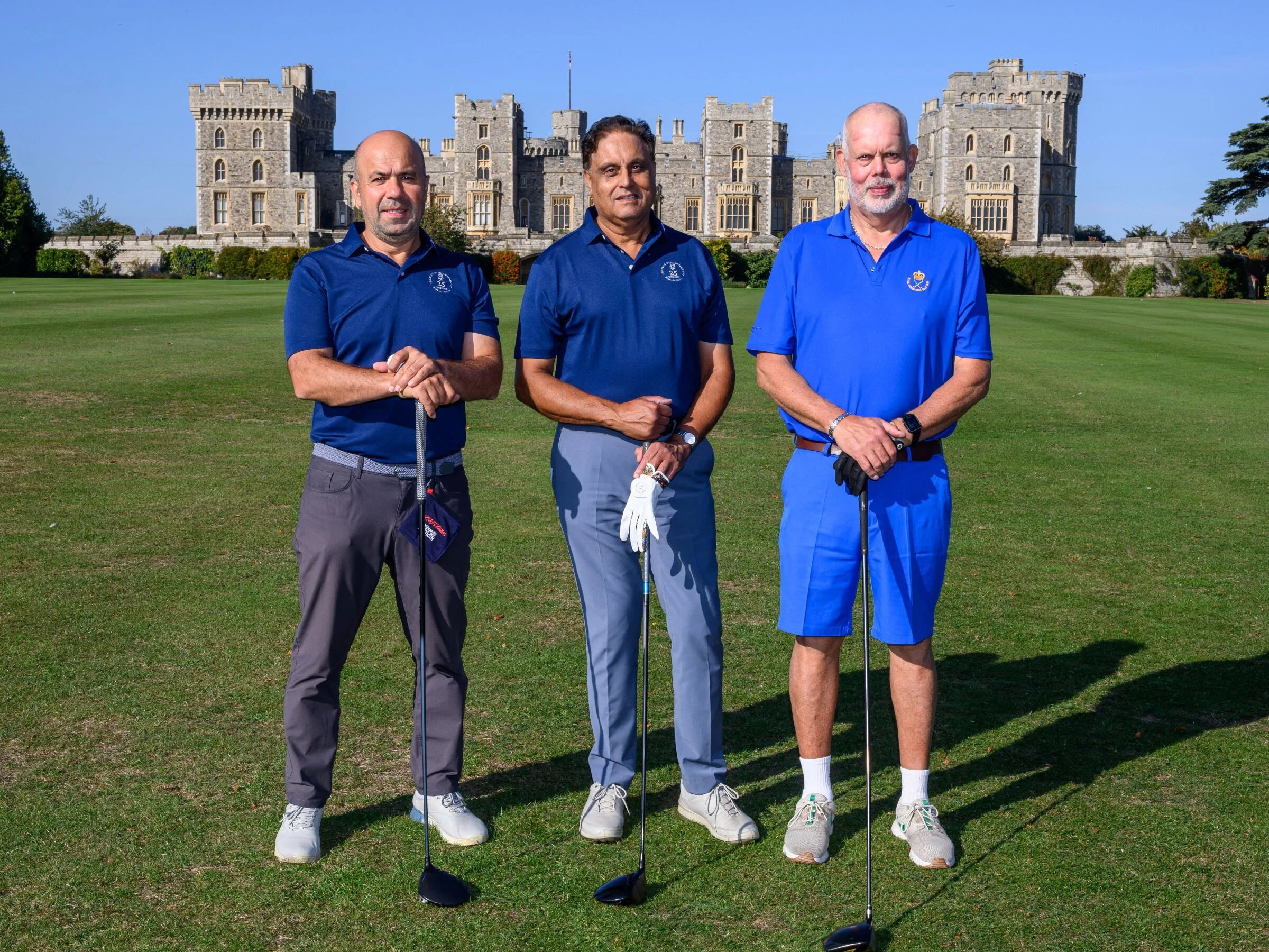 Three men standing on a golf course in front of a castle with green grass and a clear blue sky.