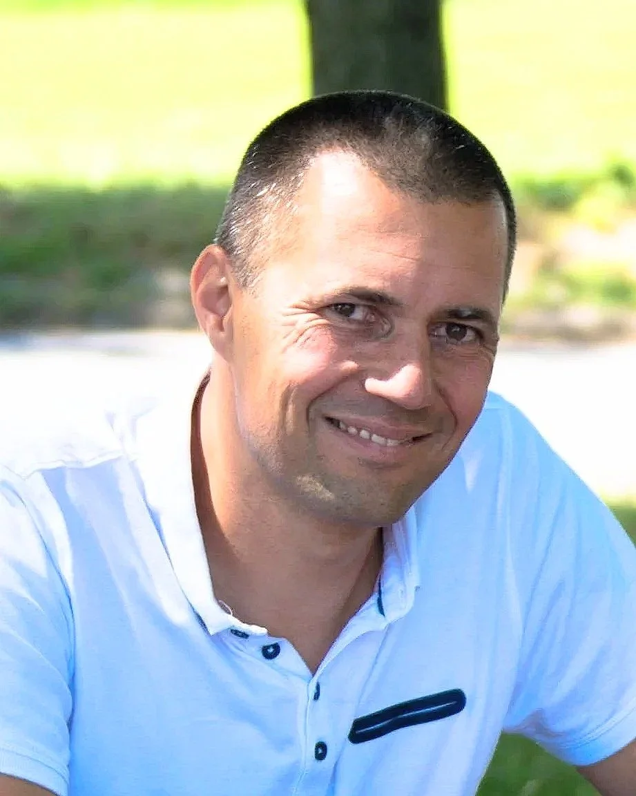 A smiling man with short dark hair wearing a white shirt outdoors on a sunny day.