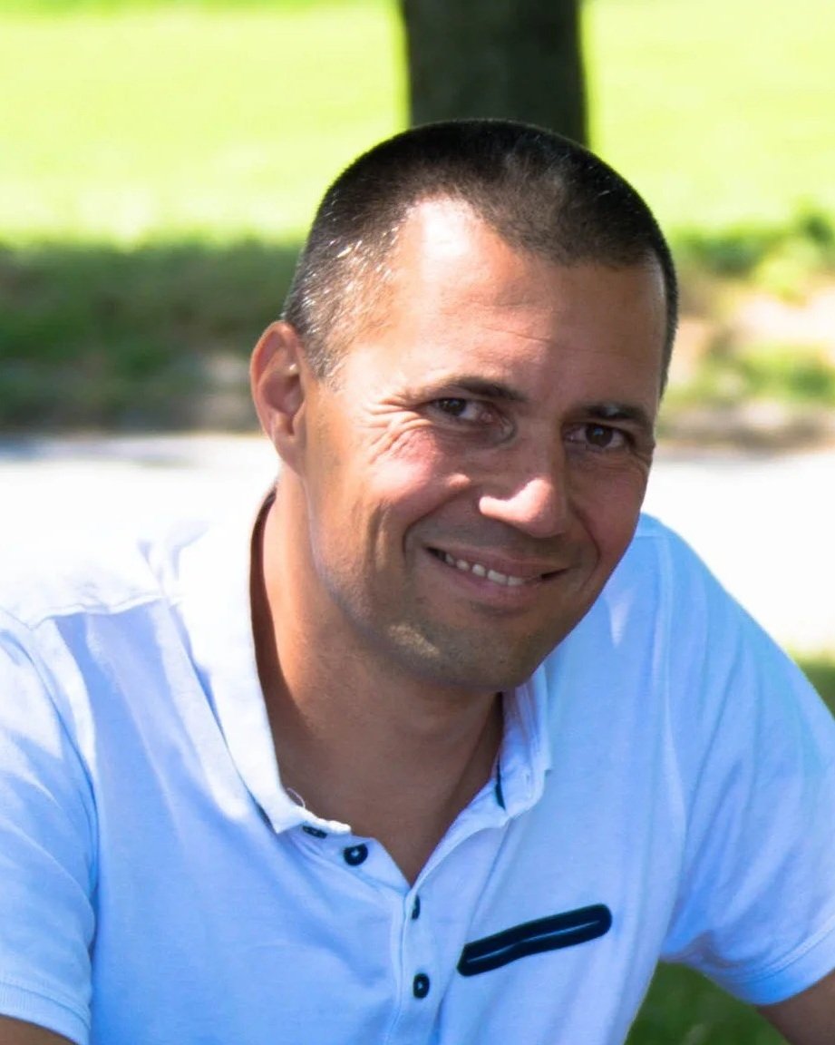 A man with short dark hair wearing a white collared shirt, smiling outdoors in a grassy area with trees in the background.