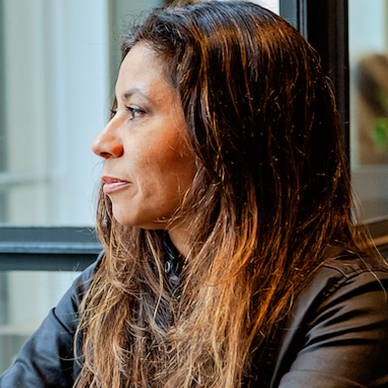 A woman with long, wavy brown hair looking out a window, sitting indoors.