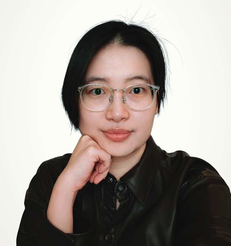 Portrait of a woman with short black hair, wearing glasses and a black leather shirt, resting her chin on her hand against a plain light background.