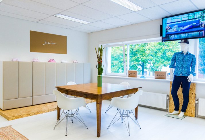 Office break room with a wooden table, four white chairs, a potted plant, a gray storage cabinet, a window with double curtains, a mannequin in a blue shirt and dark pants, and a wall-mounted TV.