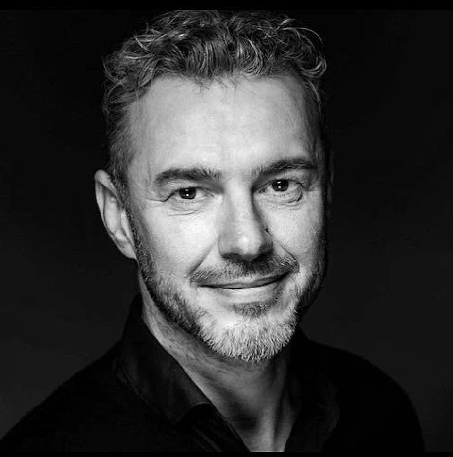 Black and white portrait of a smiling man with short, curly hair and a beard, wearing a dark shirt against a dark background.