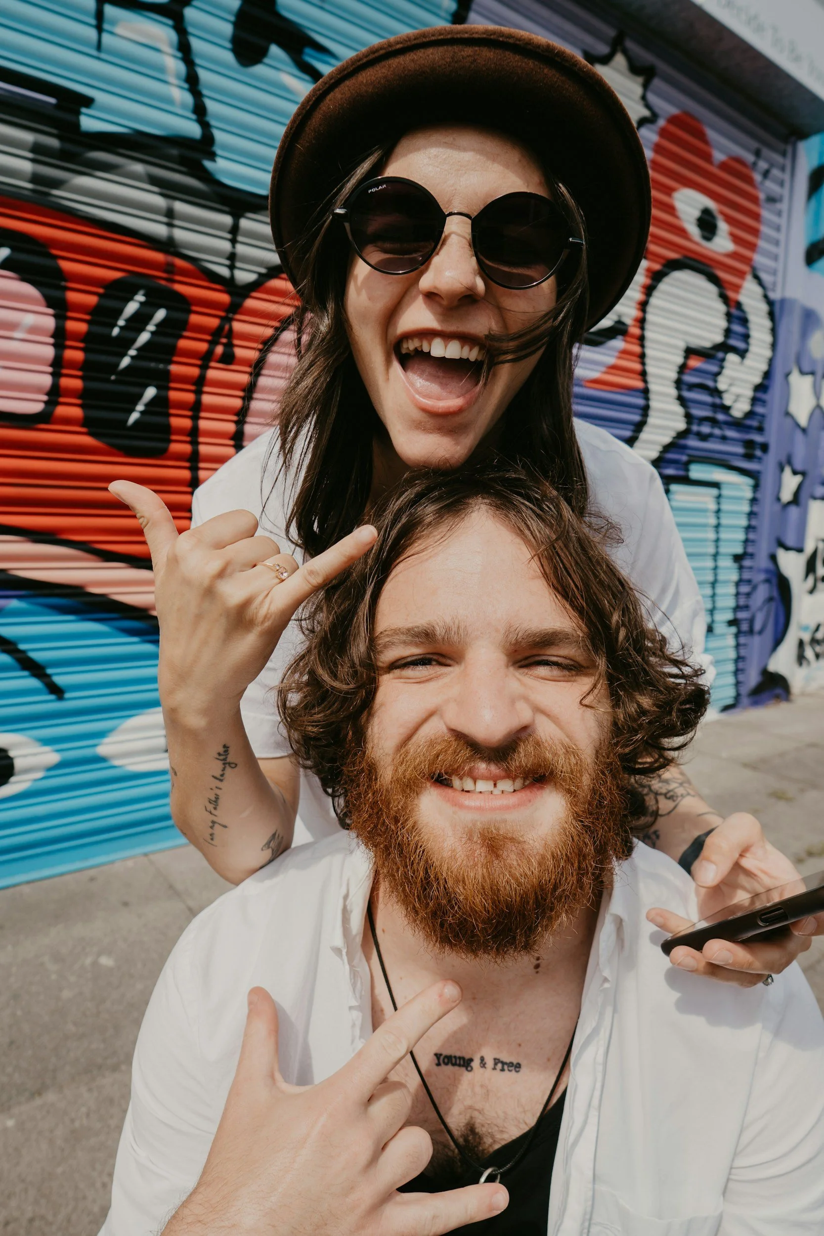 Two friends, a woman with sunglasses and a hat, and a man with a beard, smiling and making rock sign gestures in front of a colorful graffiti wall.