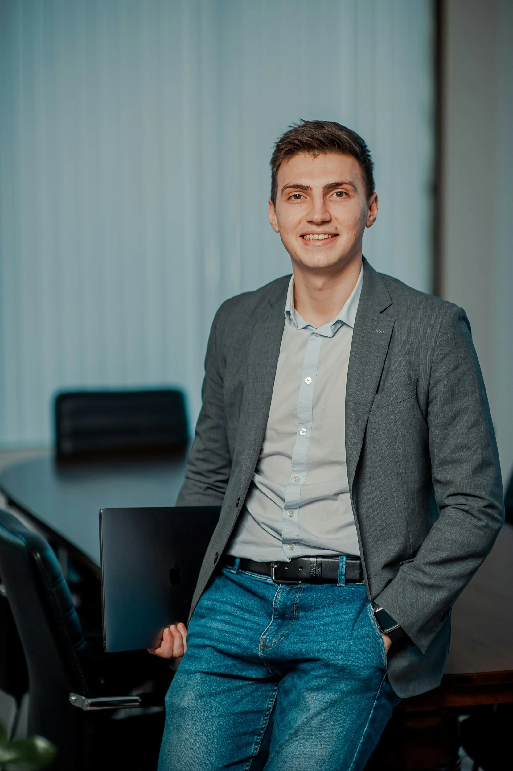 A young man in a gray blazer, light blue shirt, and jeans, holding a laptop, standing in a conference room.