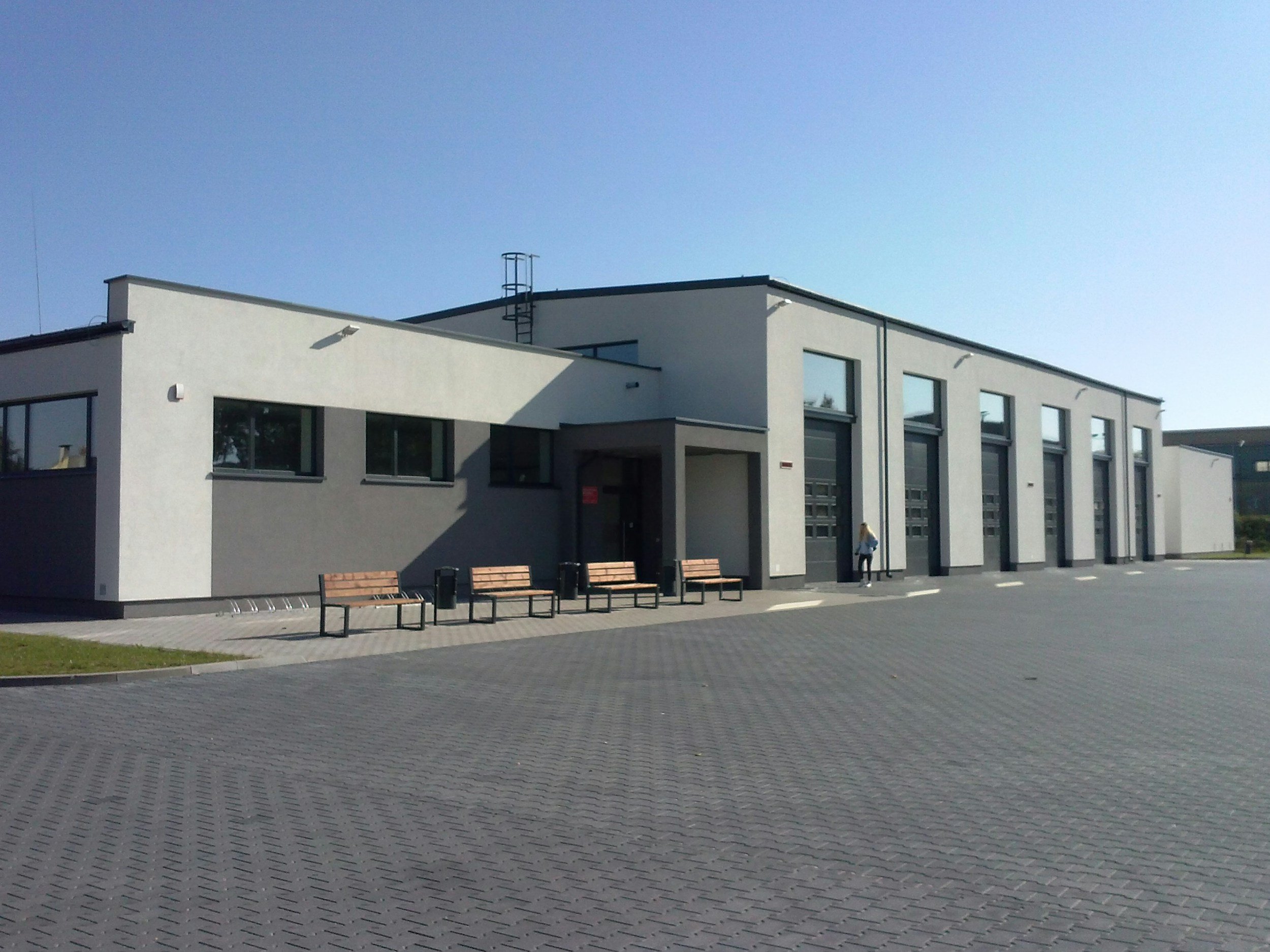 Modern white and dark gray commercial or industrial building with multiple garage doors and benches outside. Clear sky and paved area in front. warehouse clean, business clean