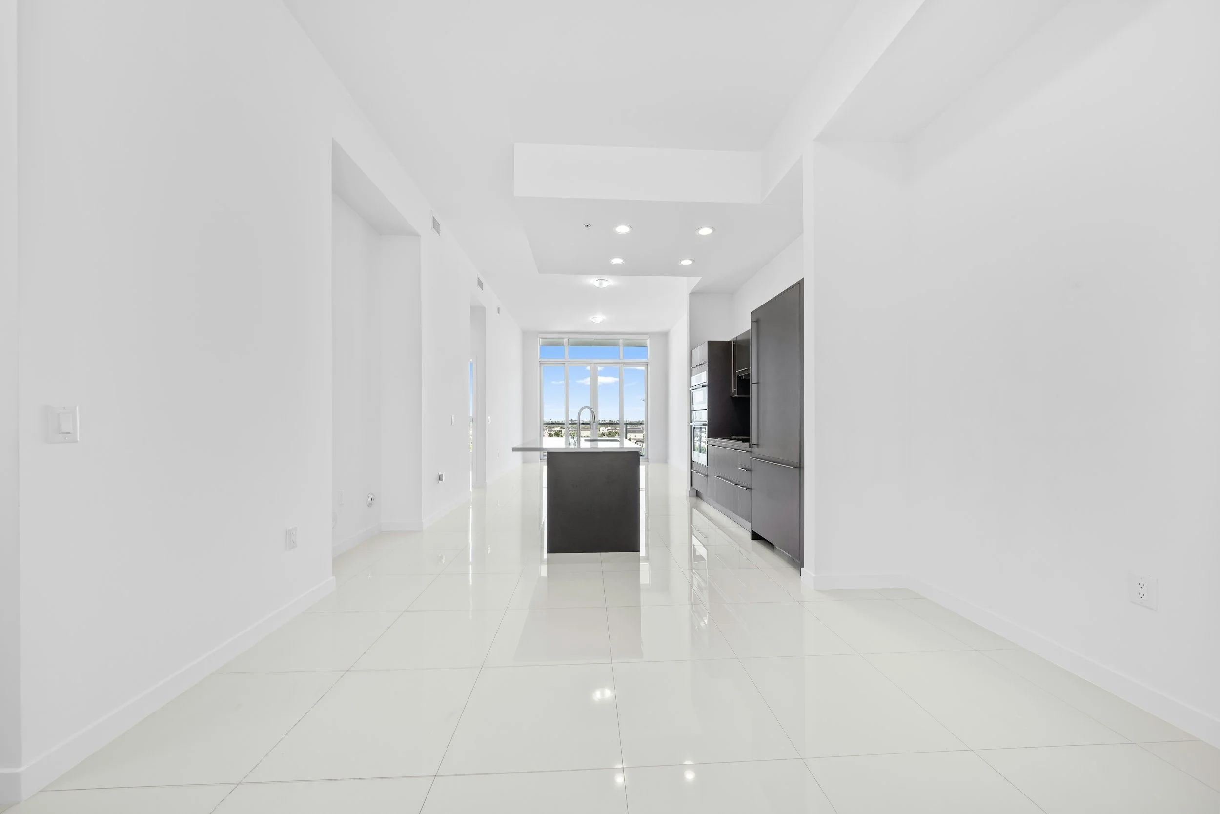 Empty modern kitchen with glossy white floor tiles, white walls, black kitchen cabinets, and a large window showing a cityscape and blue sky.