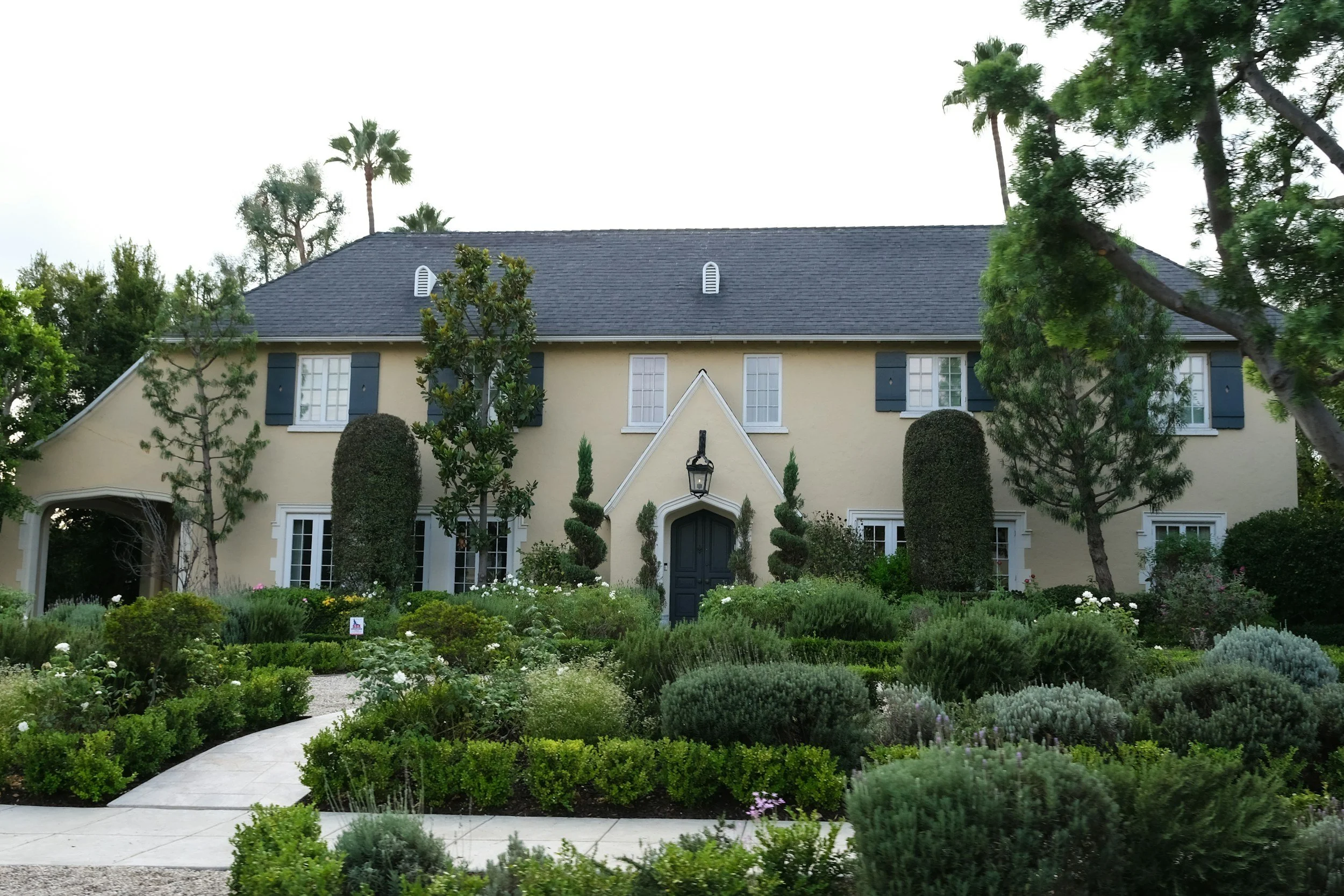A large, two-story beige house with a dark gray roof, black shutters, and a decorative front door framed by greenery, surrounded by a well-maintained garden with various bushes and trees. grounds keeping, maintenance 