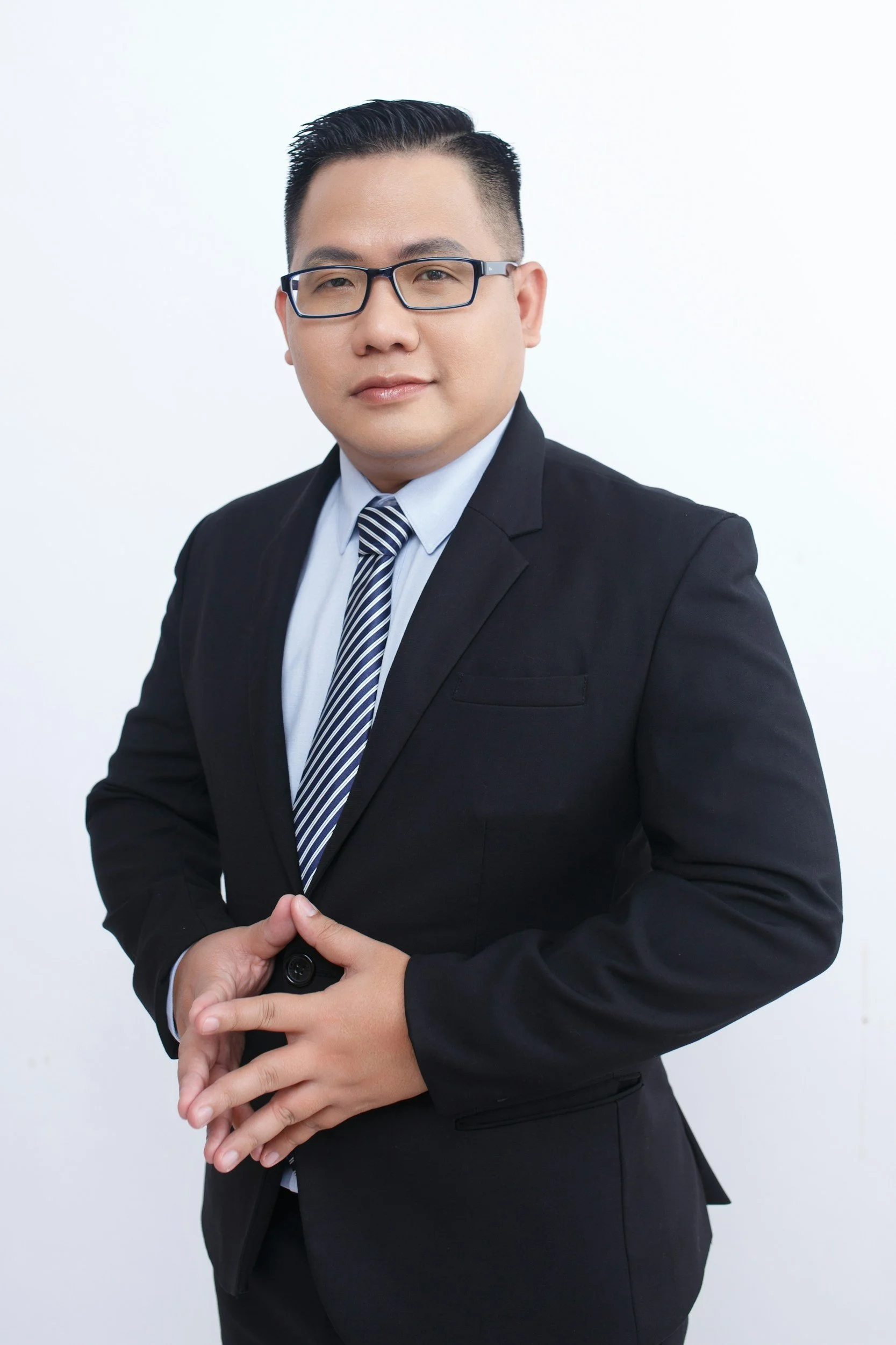 A young man in a black suit, striped tie, and glasses posing with folded hands against a white background.