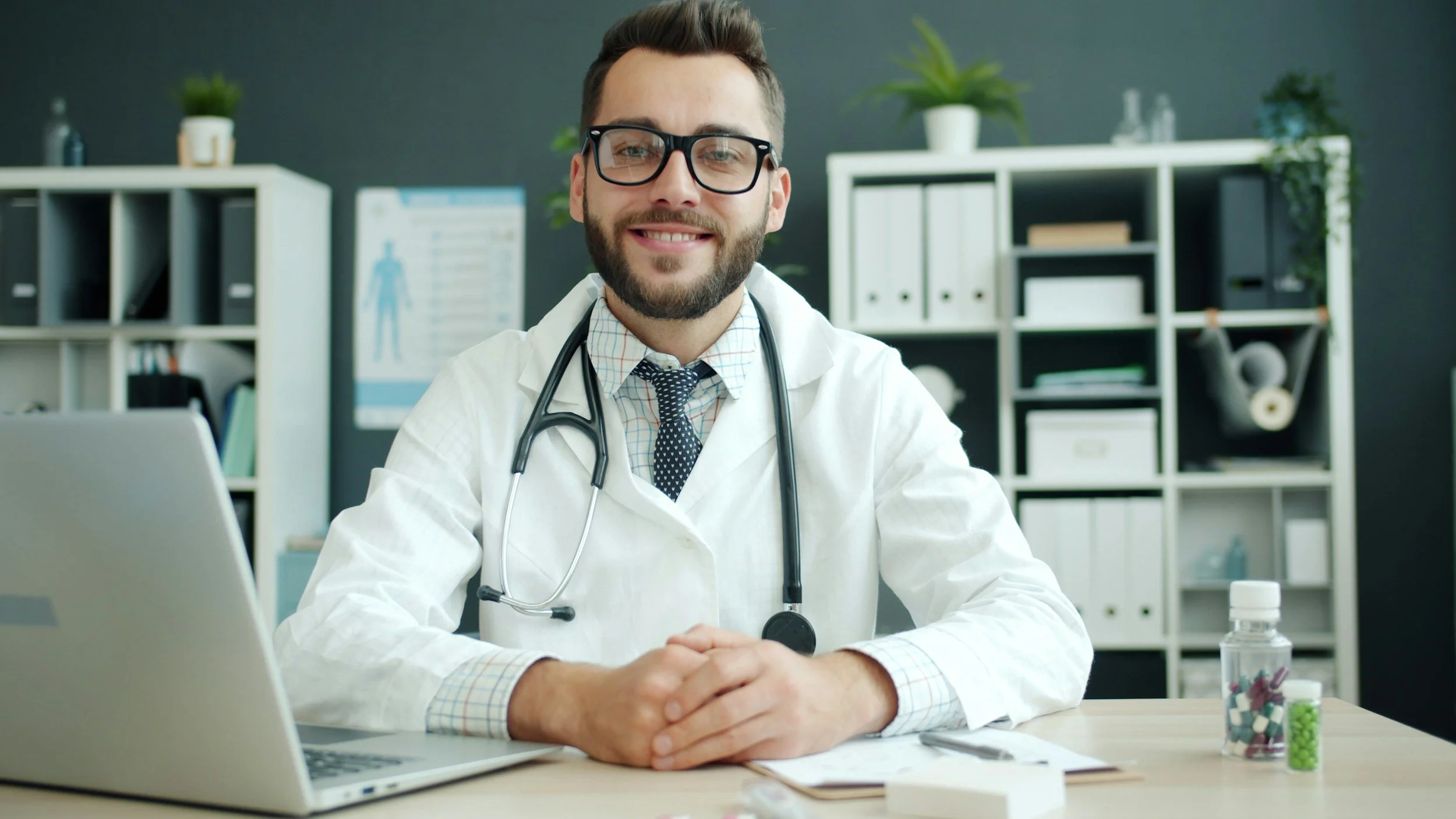 A smiling male doctor in a white coat with a stethoscope around his neck sitting at a desk in an office with shelves and medical supplies.