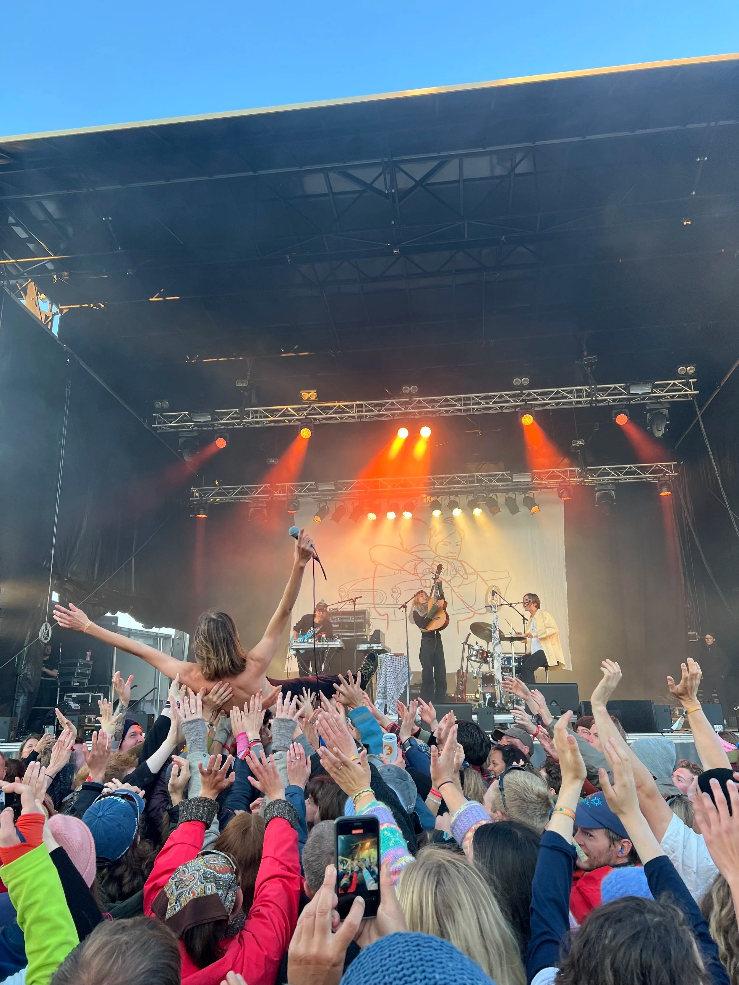 A live outdoor concert with a band performing on stage. The stage has colorful lights and a backdrop featuring a drawing. The audience is engaged, with some people raising their hands and a person crowd surfing.