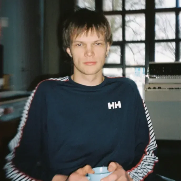 A young man with short, light brown hair, wearing a navy blue Helly Hansen sweatshirt with white, red, and black striped sleeves, sitting in a room with large windows and holding a light blue mug.