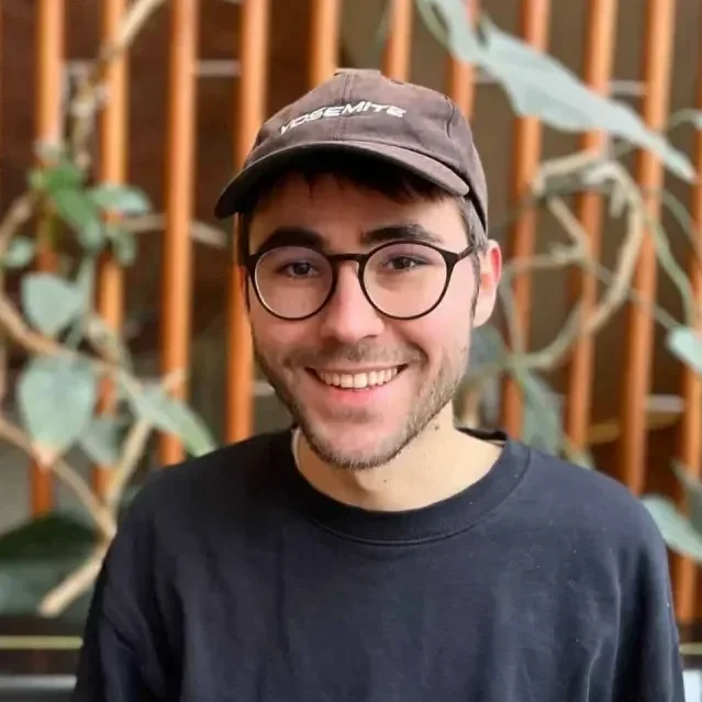 A young man with glasses, wearing a dark t-shirt and a cap, smiling outside with a wooden fence and green plants in the background.