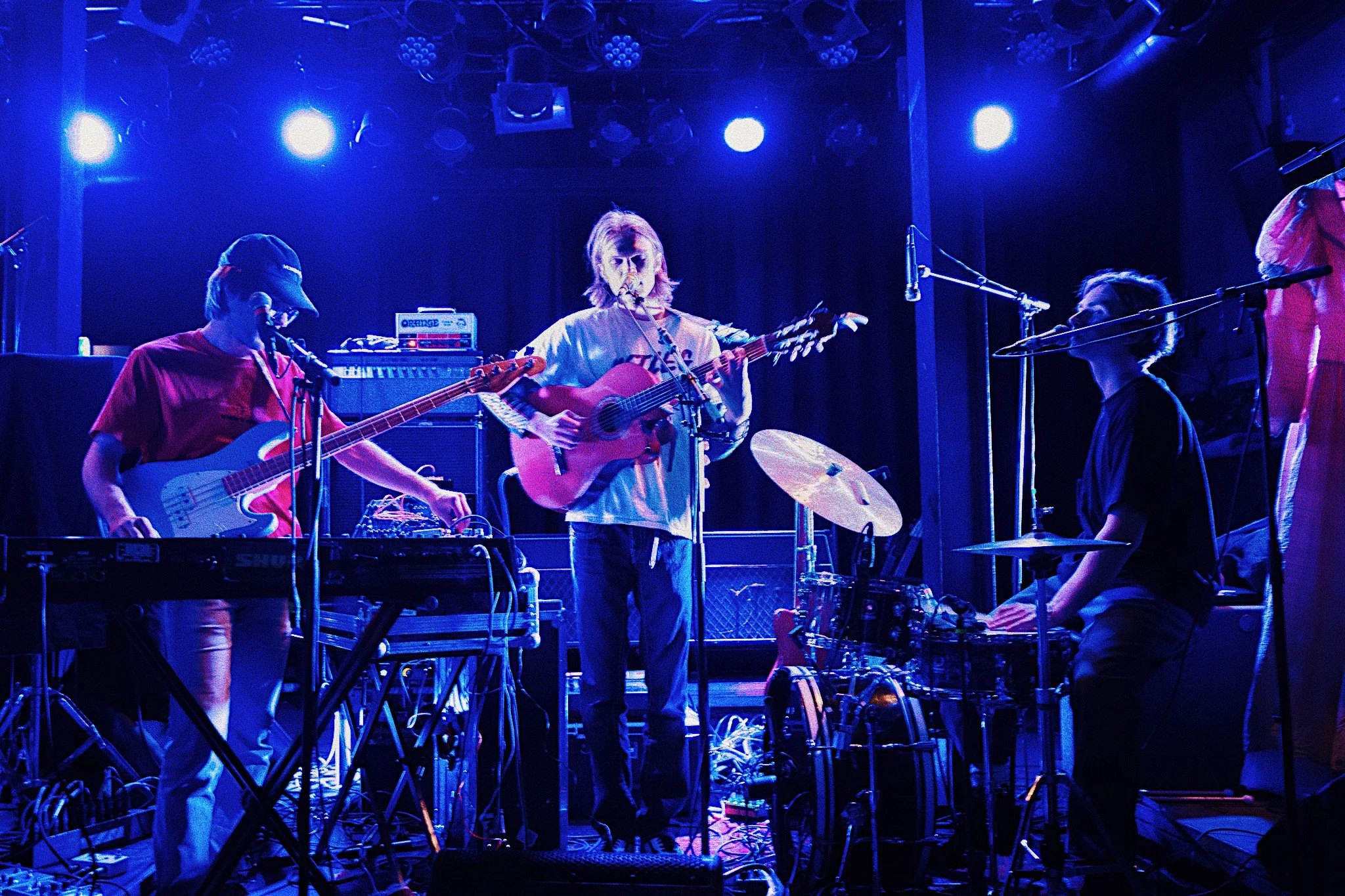 A band performs on stage with blue lighting, featuring a guitarist with long hair at center, a bassist to the left, and a drummer to the right, surrounded by various musical equipment.