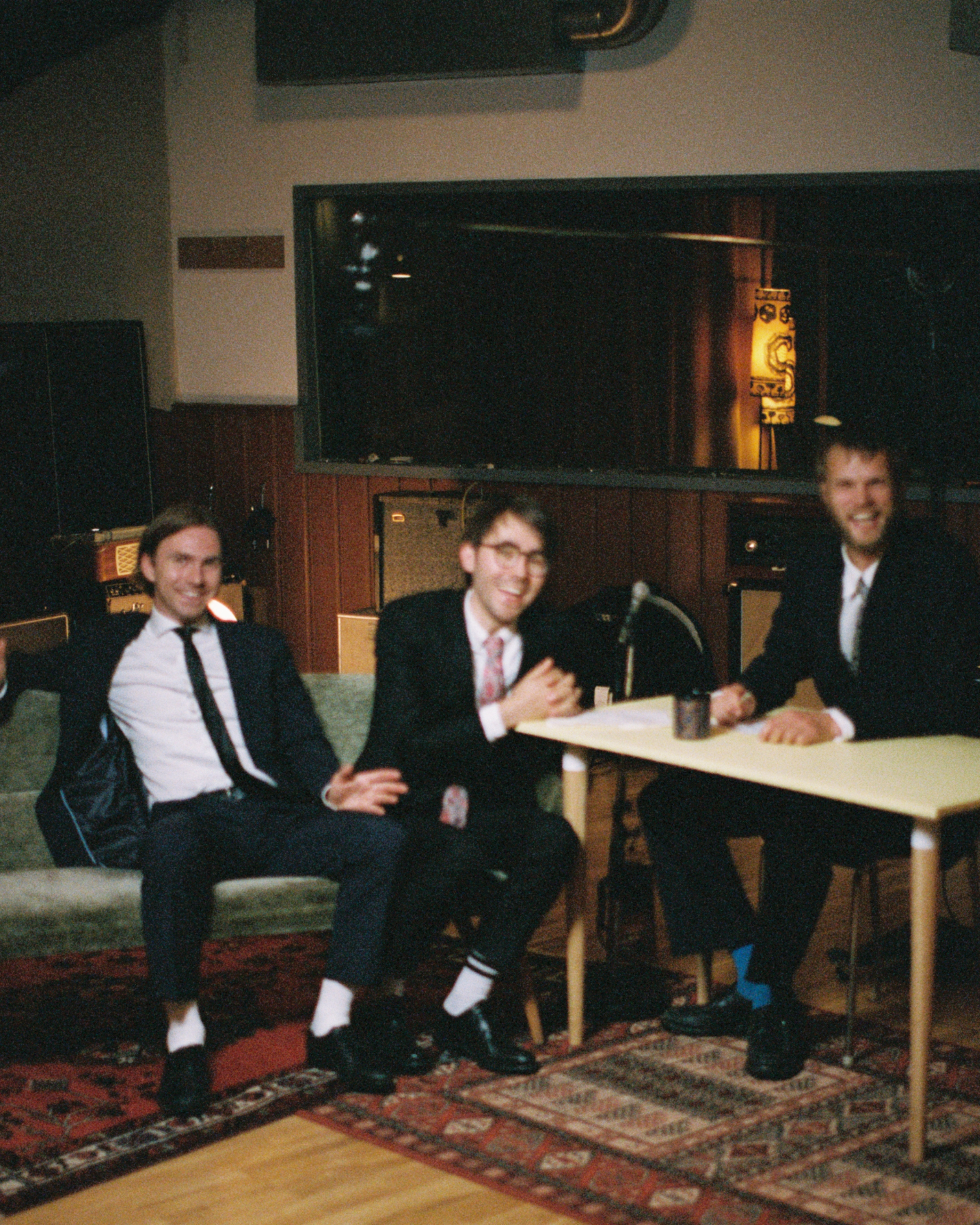 Three young men in suits sitting and smiling at a table in a dimly lit room, with a TV on the wall behind them.