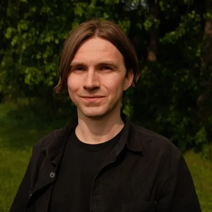 A young man with shoulder-length brown hair and fair skin, smiling slightly, wearing a black shirt against a background of green trees and grass.