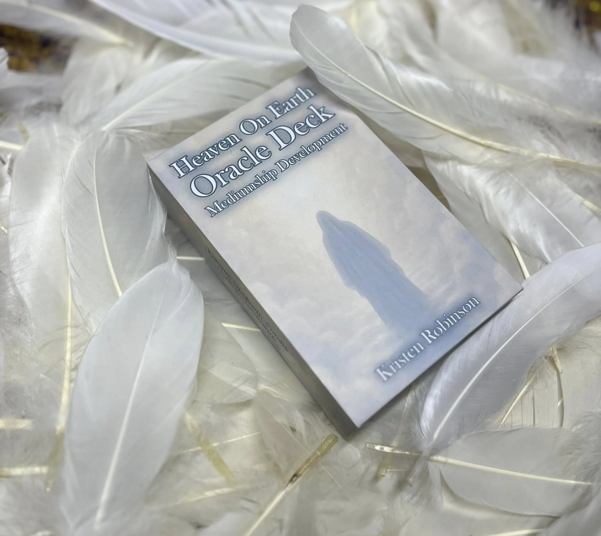 Book titled 'Heaven On Earth Oracle Deck' by Kirsten Robinson resting on a bed of white feathers.