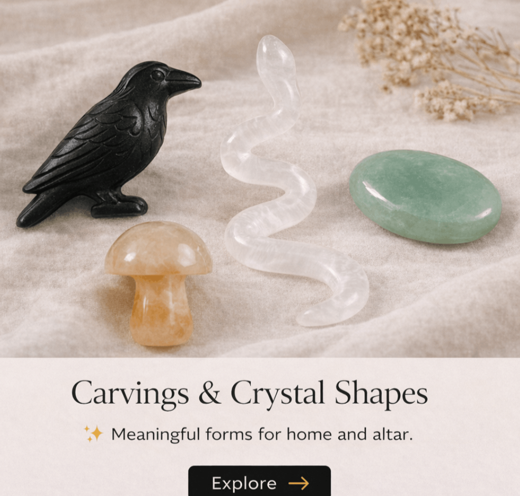 A collection of carved and crystal shapes including a black bird figure, a beige mushroom-shaped crystal, a transparent wavy crystal, and a smooth green oval-shaped stone, displayed on a neutral fabric background with some dried flowers in the corner