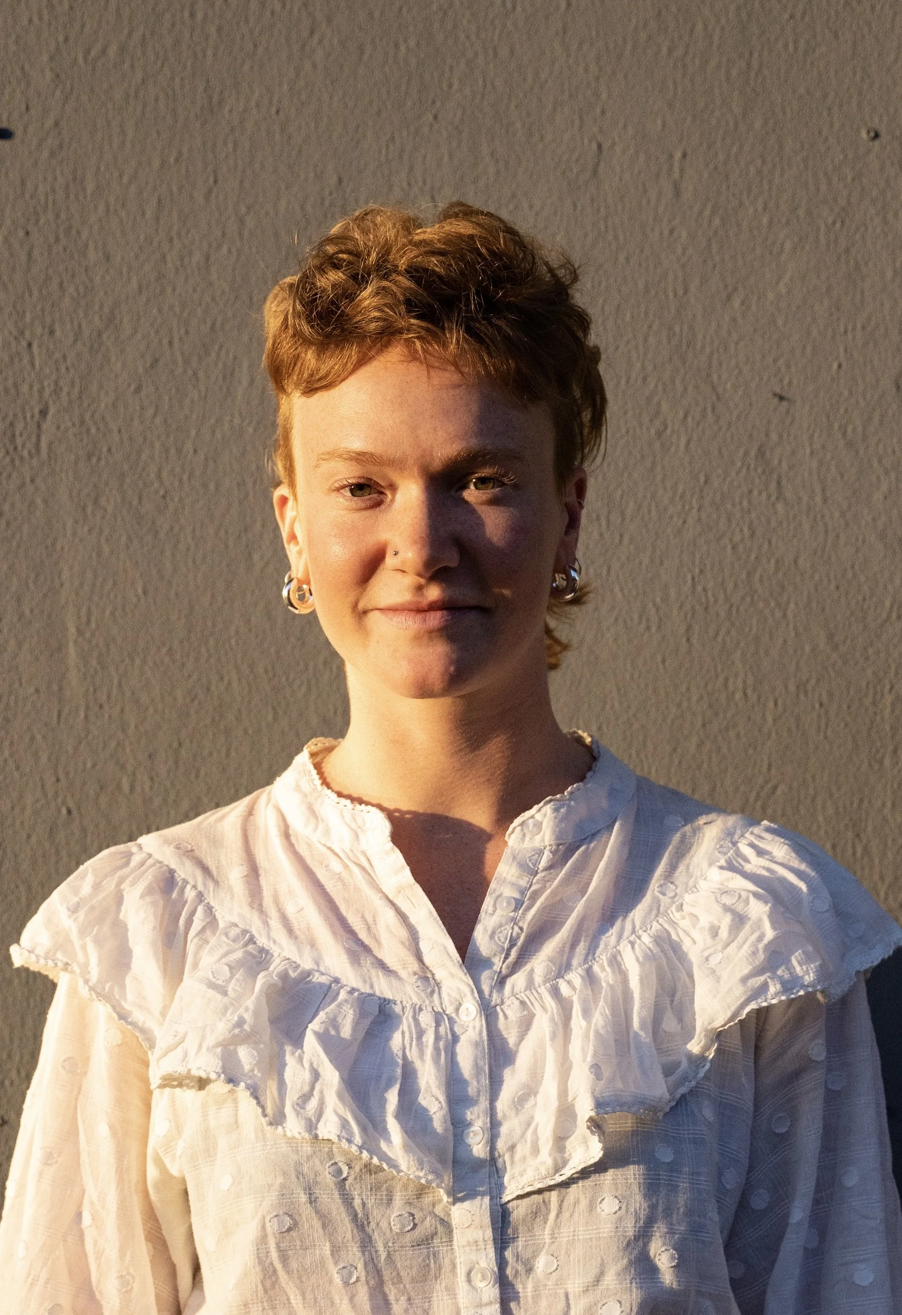 Porträt einer jungen Person mit roten Haaren, die eine weiße Bluse trägt und vor einer hellen Wand steht.