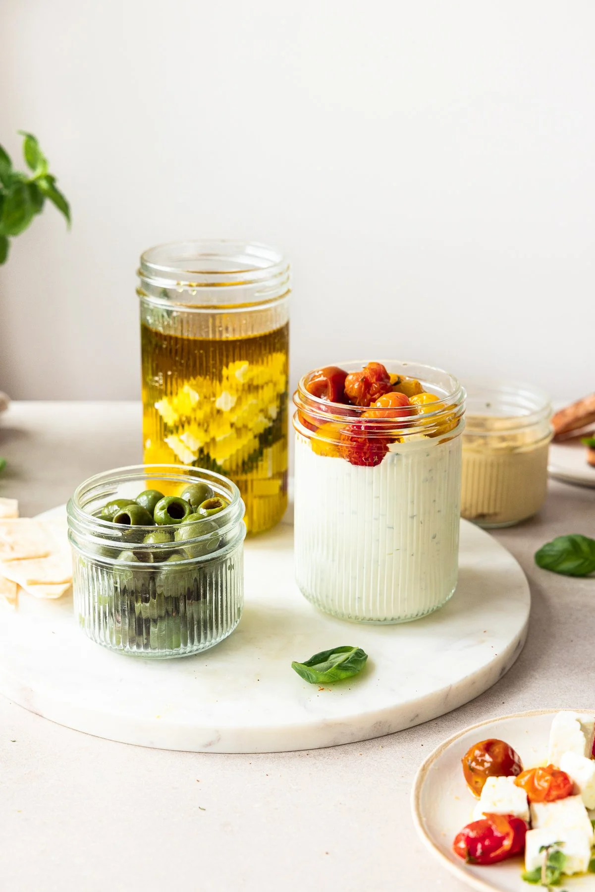 Drei Gläser mit verschiedenen Inhalten: gefüllte Oliven, eingelegte Tomaten auf Frischkäse und Öl mit Käsewürfeln. Im Hintergrund ein viertes Glas und Basilikumblätter auf einem Tisch.