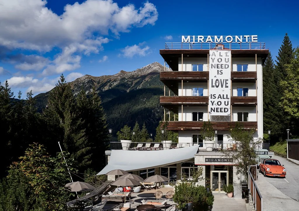 Ein Gebäude mit der Aufschrift 'Miramonte' auf dem Dach, umgeben von Bäumen und Bergen. Auf der Fassade hängt ein Banner mit dem Text 'All You Need Is Love Is All You Need'. Davor befindet sich eine Terrasse mit Tischen und Sonnenschirmen und ein rotes Auto ist geparkt.