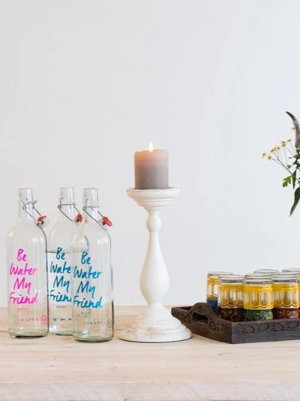 Ein Tisch mit drei klaren Glasflaschen mit der Aufschrift "Be Water My Friend", einem weißen Kerzenhalter mit einer brennenden Kerze, einem Tablett mit mehreren Gewürzgläsern und Blumen im Hintergrund.