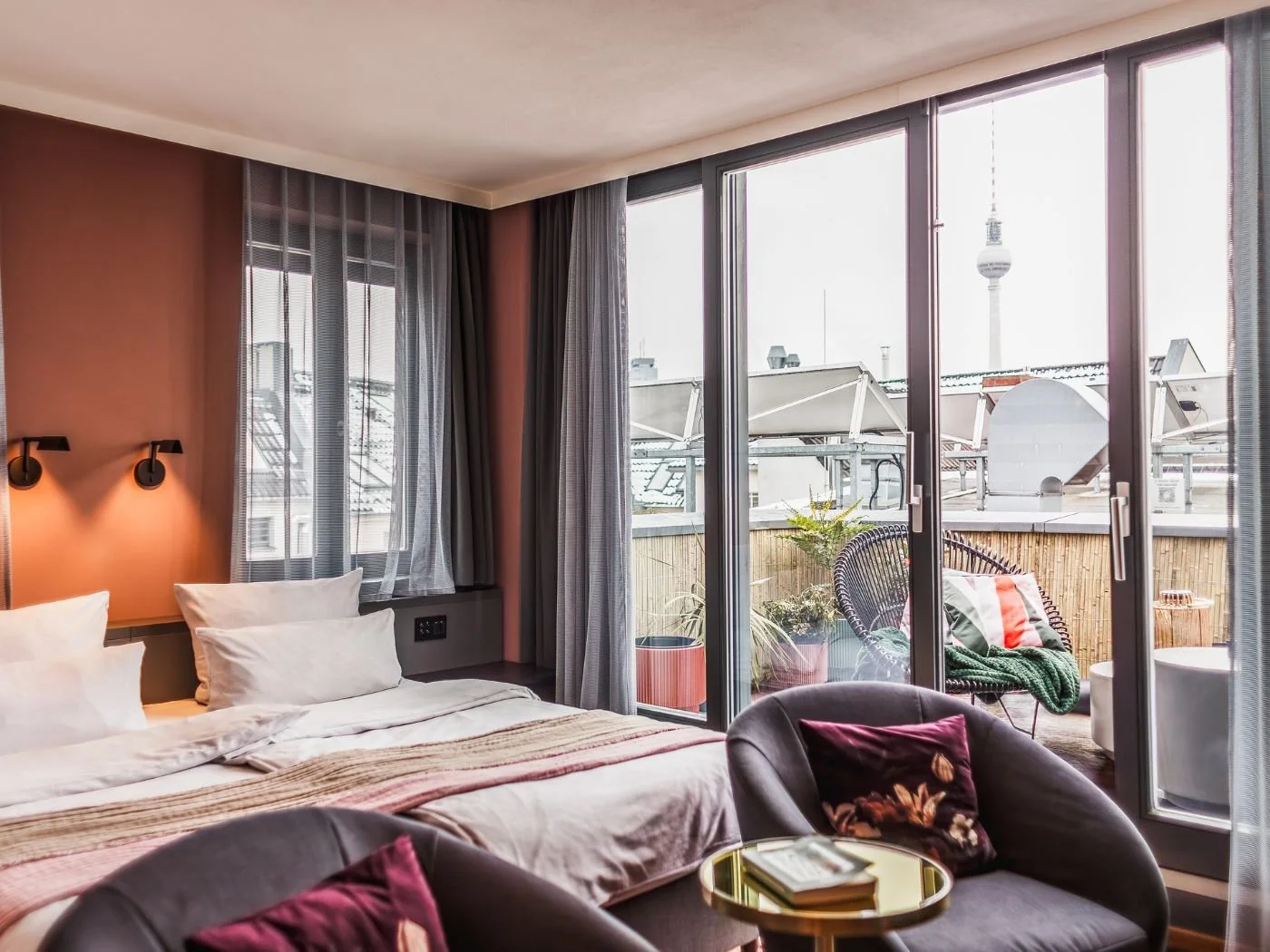 Modernes Hotelzimmer mit Doppelbett, bequemen Sesseln und Tisch, Fensterfront mit Blick auf eine Terrasse und den Berliner Fernsehturm im Hintergrund.