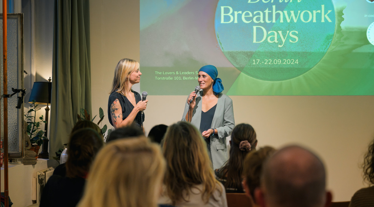 Zwei Frauen halten Mikrofone und sprechen vor einem Publikum. Im Hintergrund ist ein Projektorbild mit der Aufschrift 'Berlin Breathwork Days' und Veranstaltungsdetails.