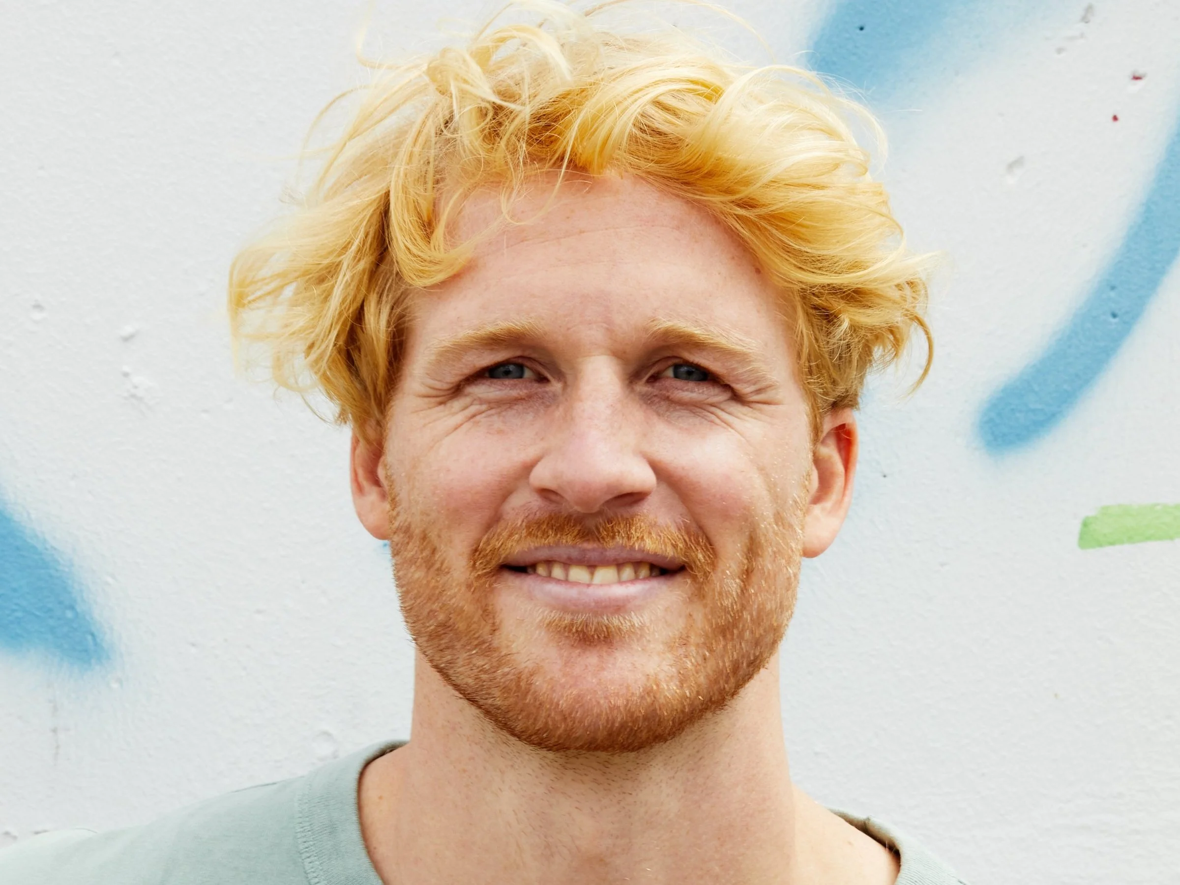 Nahaufnahme eines lachenden Mannes mit roten Haaren, rotem Bart und blauen Augen vor einer weißen Wand mit bunten Sprühfarben