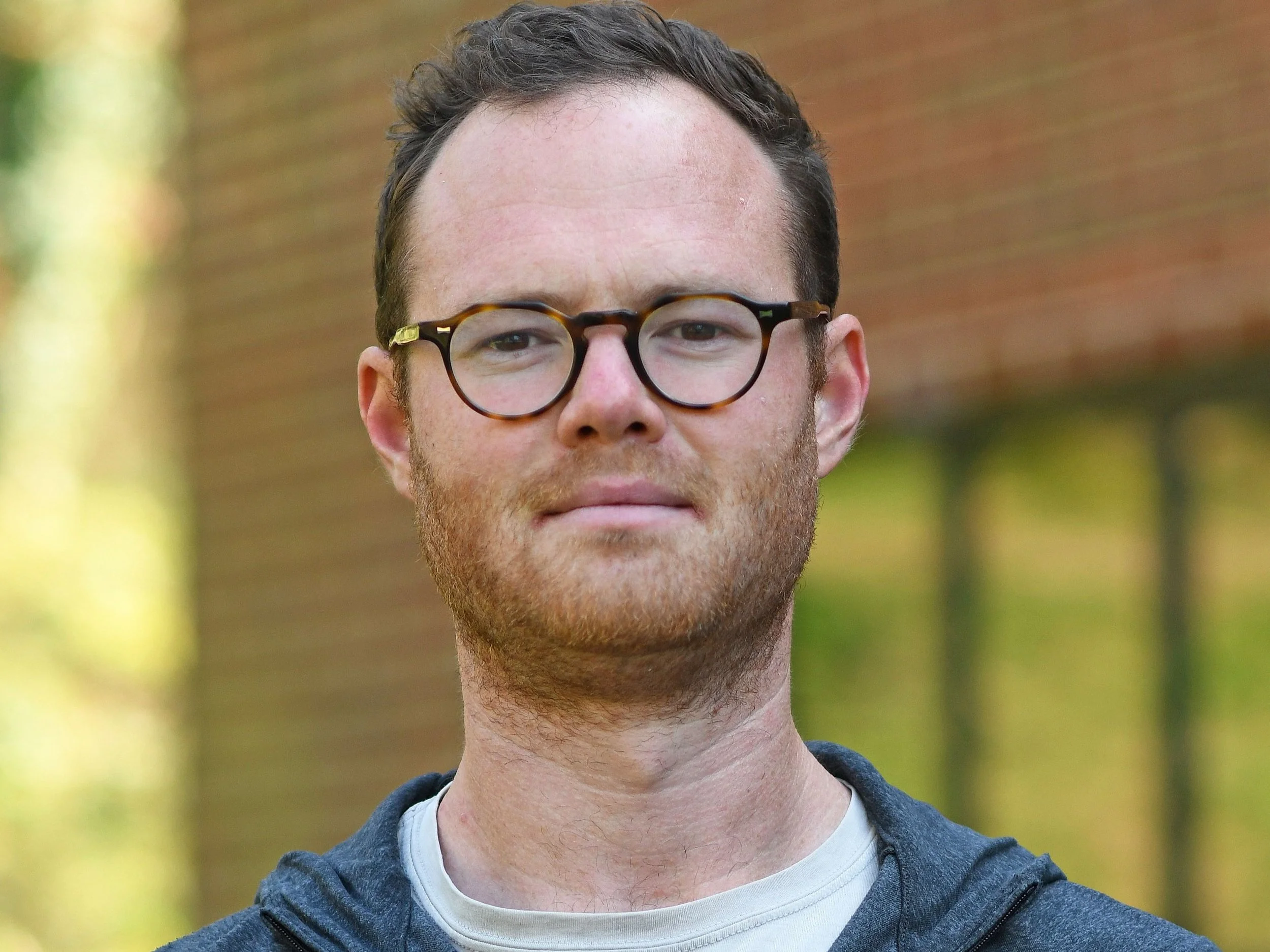 Junge Mann mit Brille, kurze braune Haare, im Freien vor einem Baum. Er trägt ein graues Hoodie und ein weißes T-Shirt.