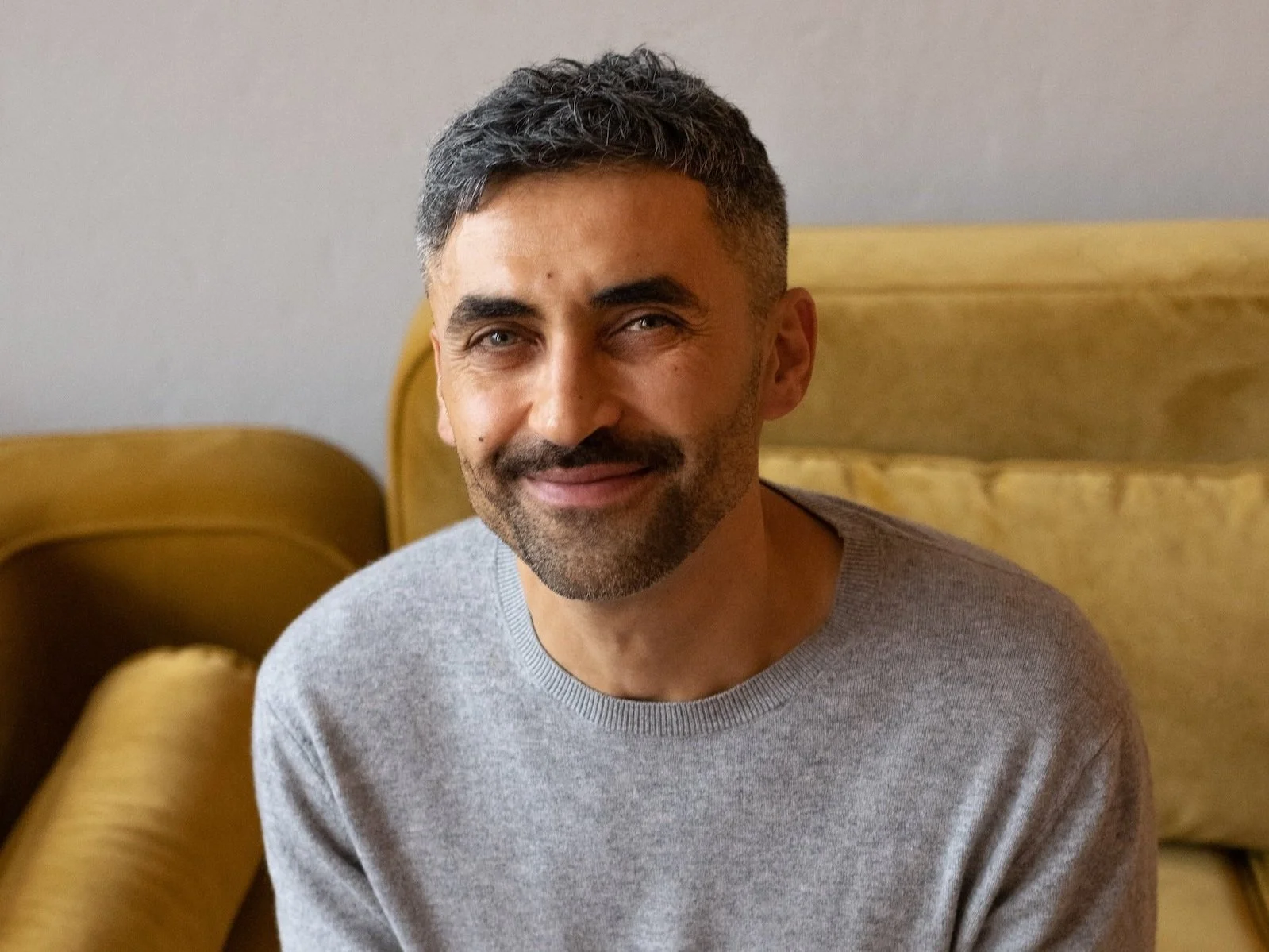 Ein lächelnder Mann mit dunklem Haar, Bart und grauem Pullover sitzt auf einem gelben Sofa.