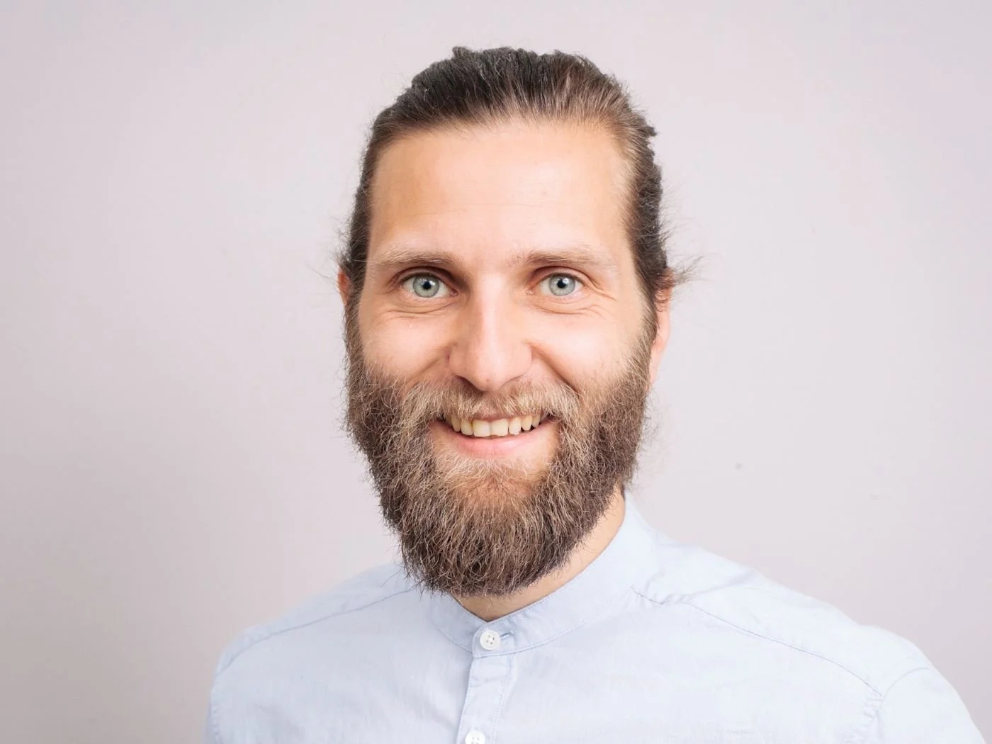 Ein lächelnder Mann mit langem Haar, Brille, voller Bart und blauen Augen vor hellem Hintergrund.