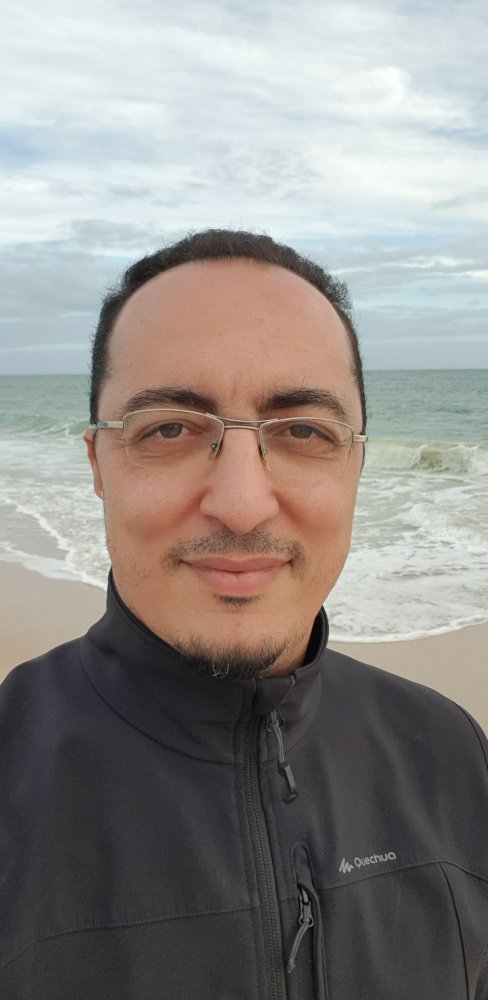 Ein Mann trägt eine schwarze Jacke mit einem Reißverschluss und steht am Strand vor dem Meer bei bewölktem Himmel.