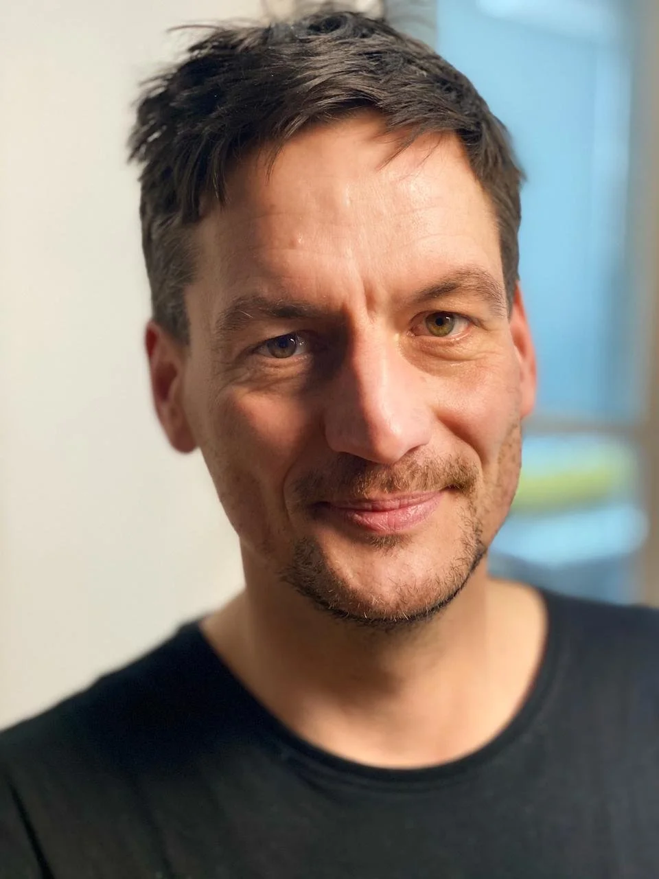 Nahaufnahme eines lächelnden Mannes mit kurzen braunen Haaren, hellen Augen, leichter Stoppelschnauze und schwarzem T-Shirt, vor einem neutralen Hintergrund.