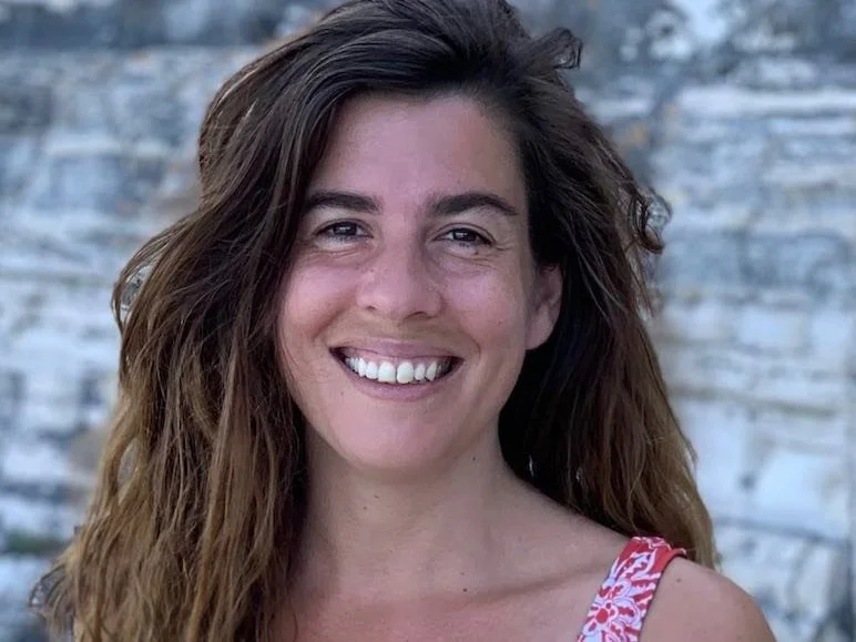 Eine junge Frau mit langen braunen Haaren, lächelnd vor einem steinernen Hintergrund.