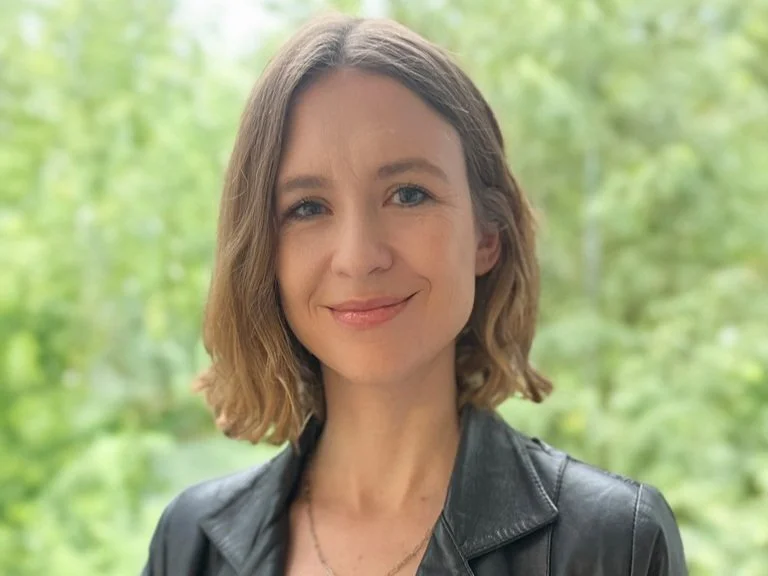 Porträt einer jungen Frau mit braunen, lockeren Haaren, lächelnd, vor einem grünen, bewaldeten Hintergrund, trägt eine schwarze Lederjacke.