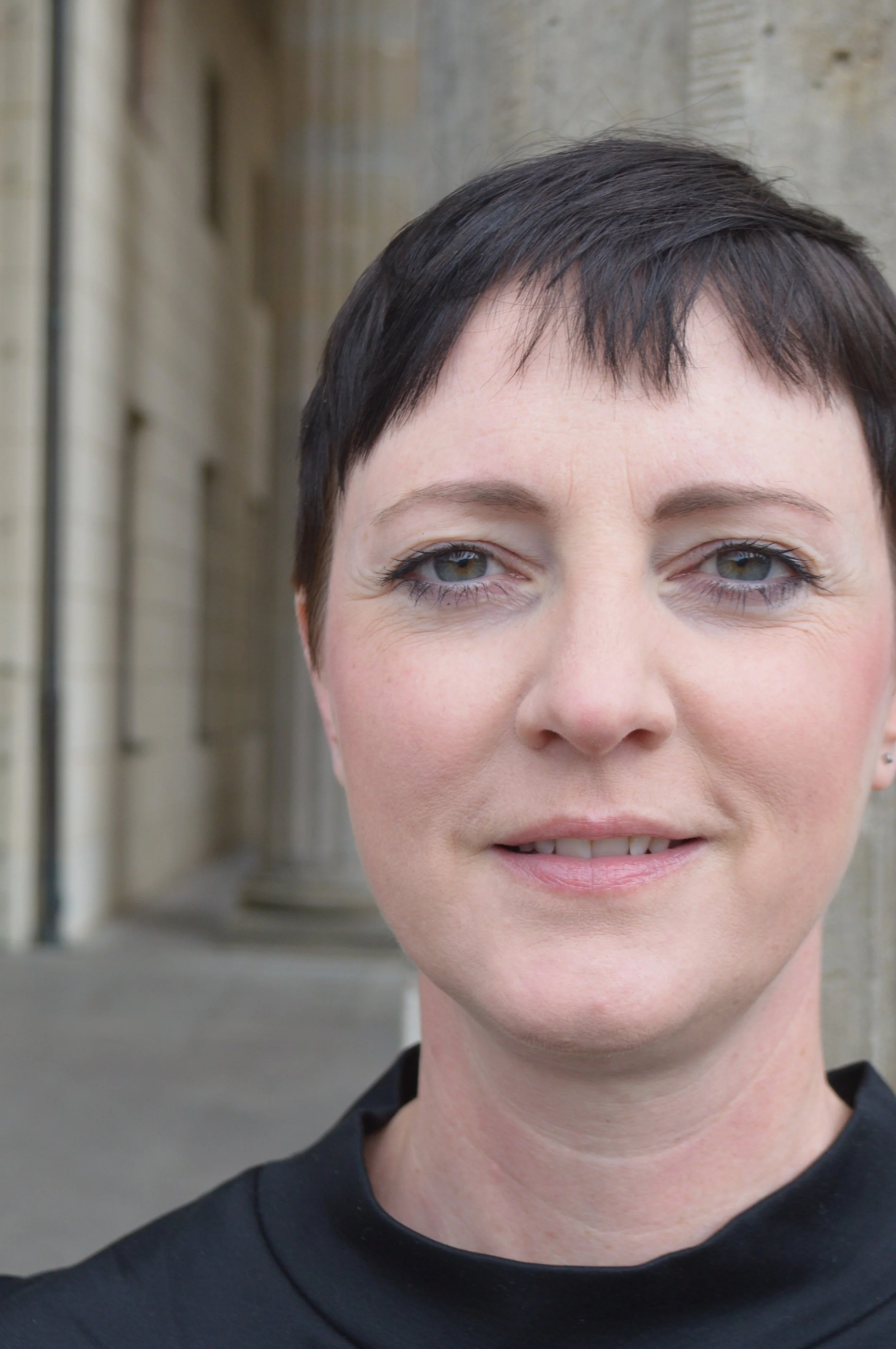 Nahaufnahme eines lächelnden Frauenkopfes vor einem steinernen Gebäude im Freien.