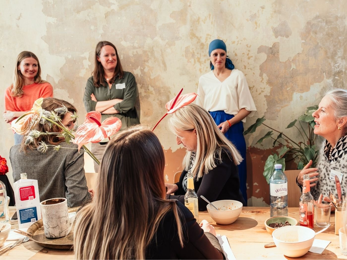Gruppe von Menschen, die an einem gemütlichen Tisch in einem Café oder Restaurant sitzen, mit Blumen auf dem Tisch. Einige Personen stehen im Hintergrund. Es herrscht eine entspannte und gesellige Atmosphäre.