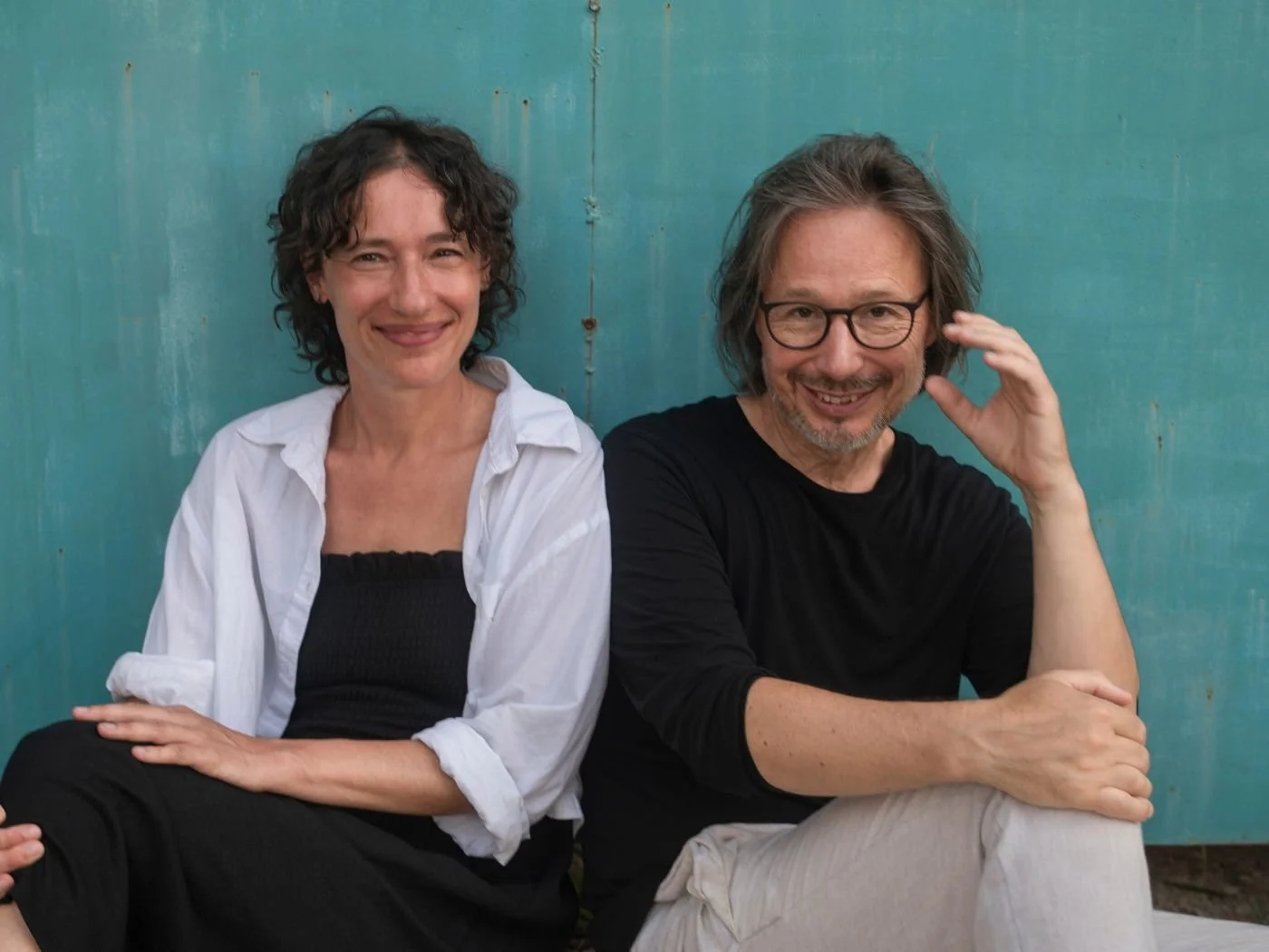 Ein Mann und eine Frau sitzen nebeneinander vor einer blauen Wand, lächeln und schauen zur Kamera.