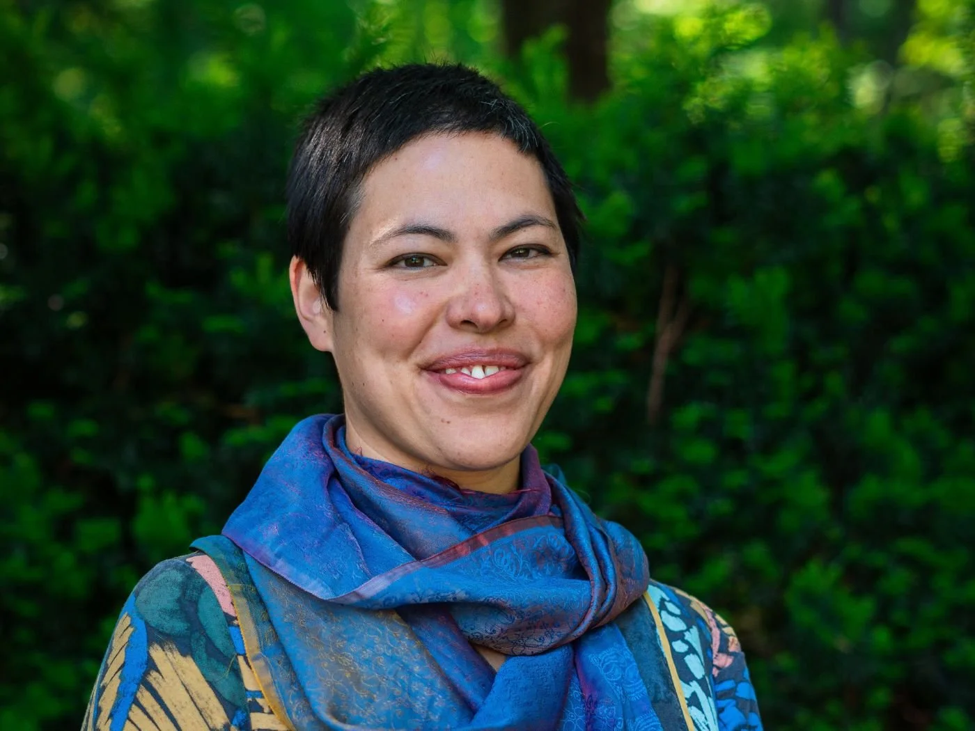 Nahaufnahme einer lächelnden Frau mit kurzen schwarzen Haaren, blasser Haut und einem blauen, bunten Schal, vor einem grünen, bewaldeten Hintergrund.