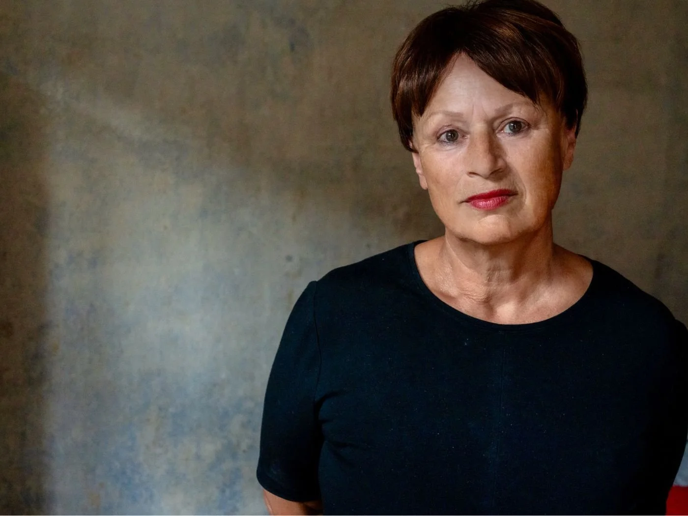 Porträt einer älteren Frau mit kurzen braunen Haaren, rotem Lippenstift und schwarzem Oberteil, vor einem neutralen Hintergrund.