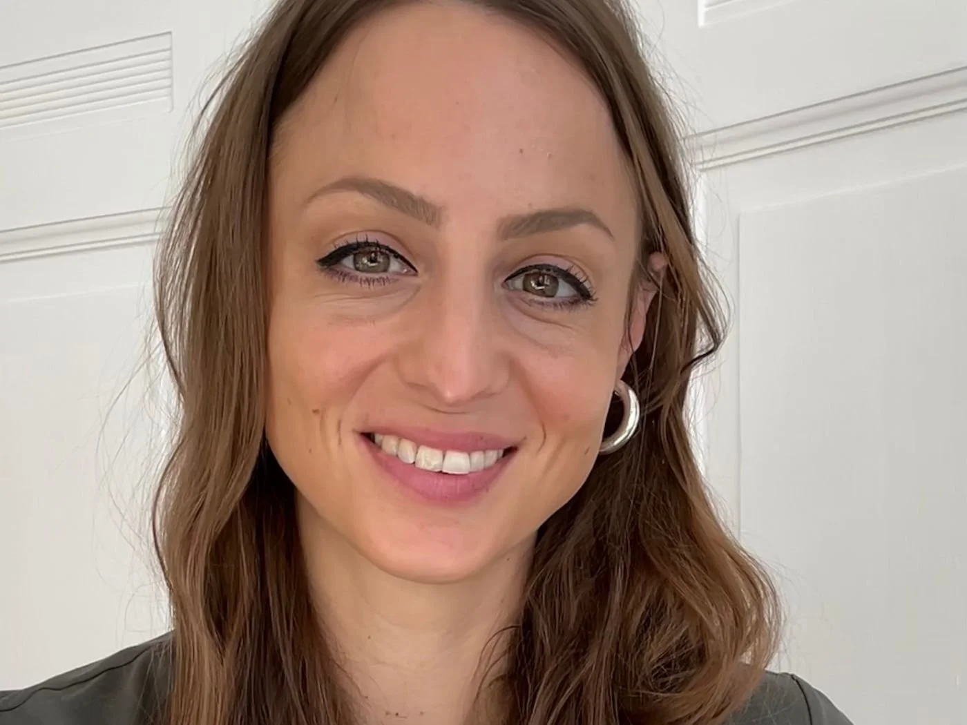 Nahaufnahme eines lachenden Frau mit braunen, gewellten Haaren, silbernen Creolen, dunklem Oberteil, vor weißer Wand.