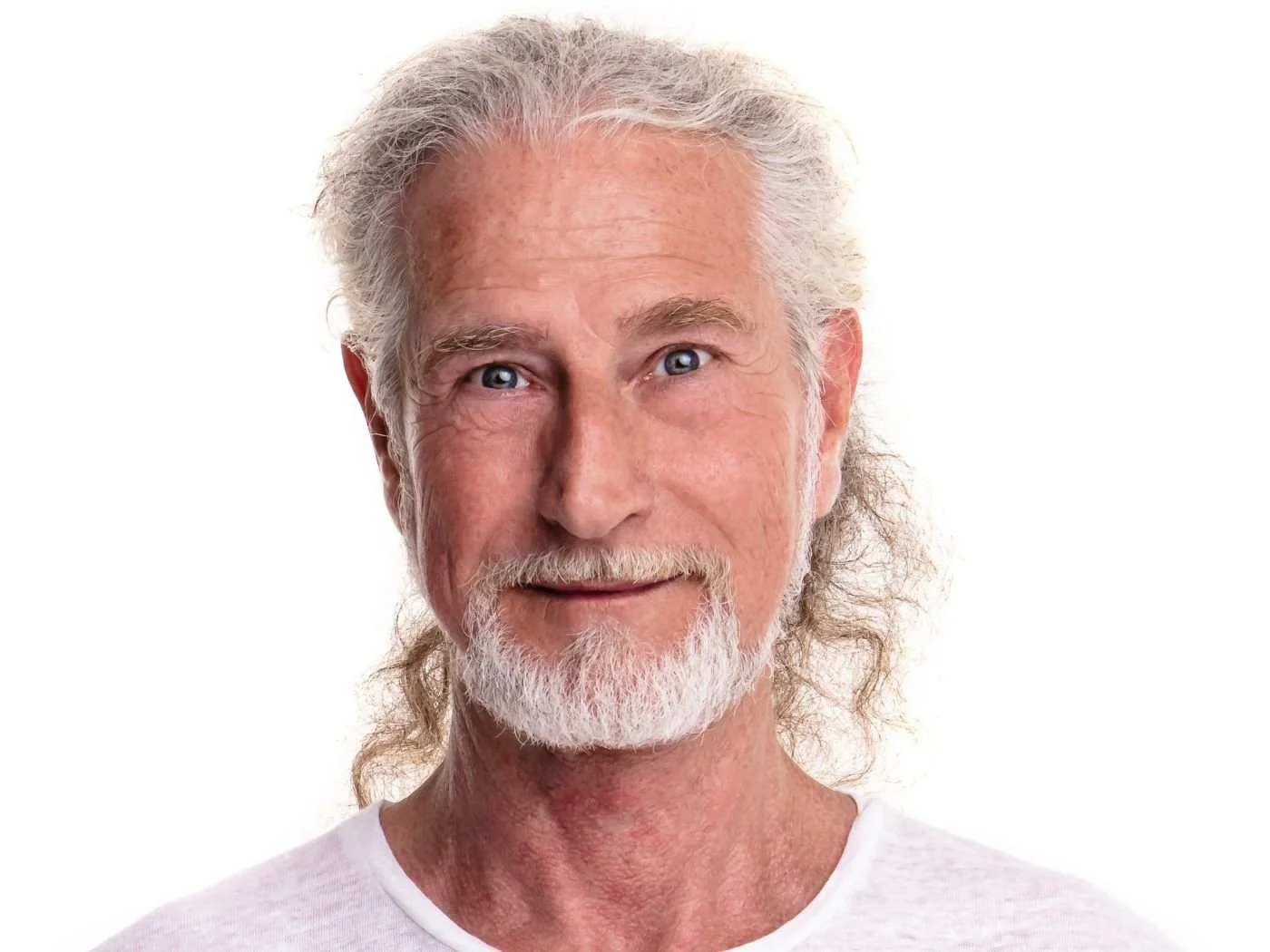 Ein älterer Mann mit langen weißen Haaren und Bart, trägt ein weißes T-Shirt, lächelt vor einem weißen Hintergrund.