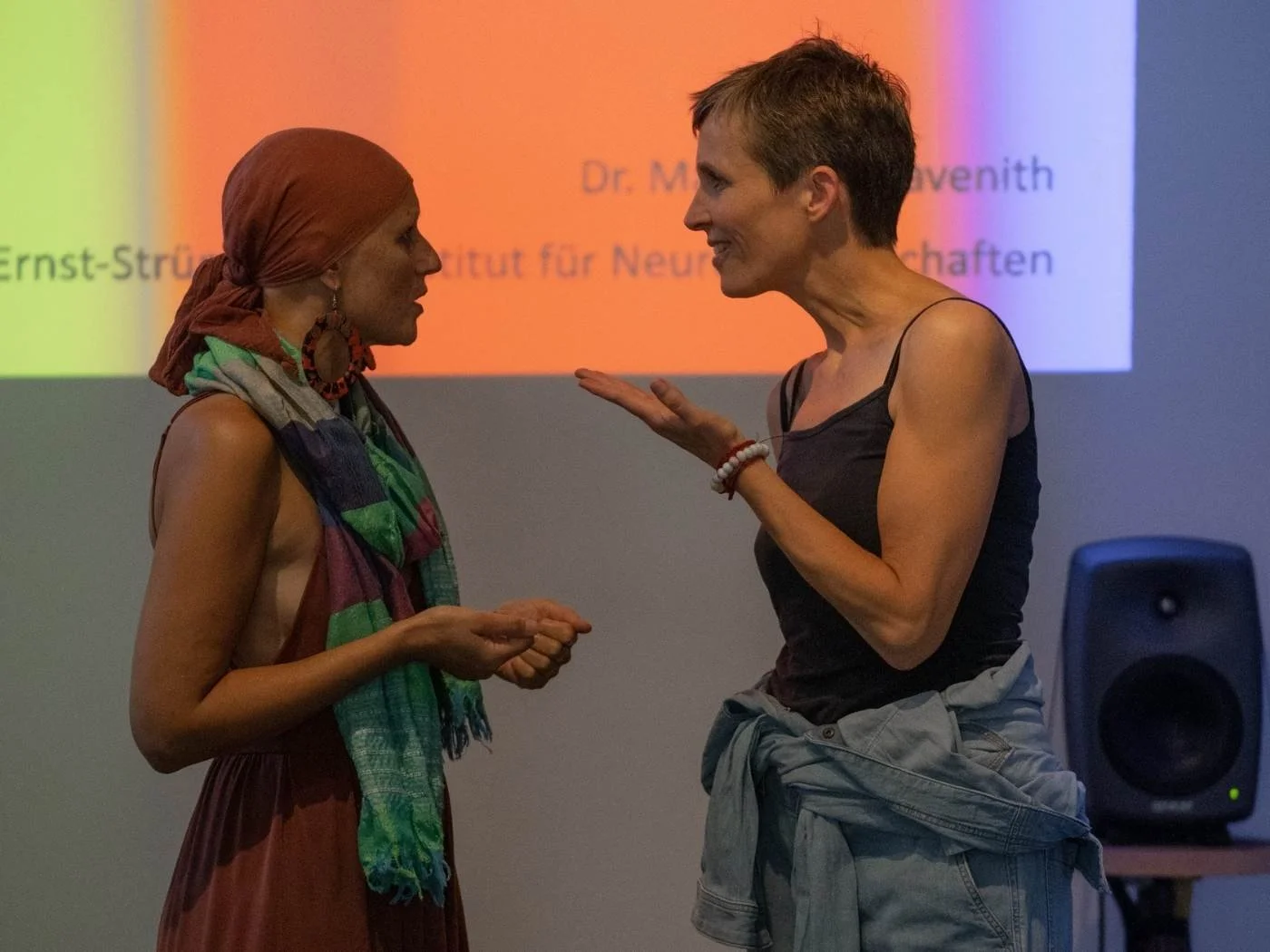 Zwei Frauen im Gespräch, eine trägt ein buntes Tuch und ein Kopftuch, die andere trägt ein schwarzes Top und eine Jeansjacke um die Taille, im Hintergrund ein Projektor und ein Lautsprecher.