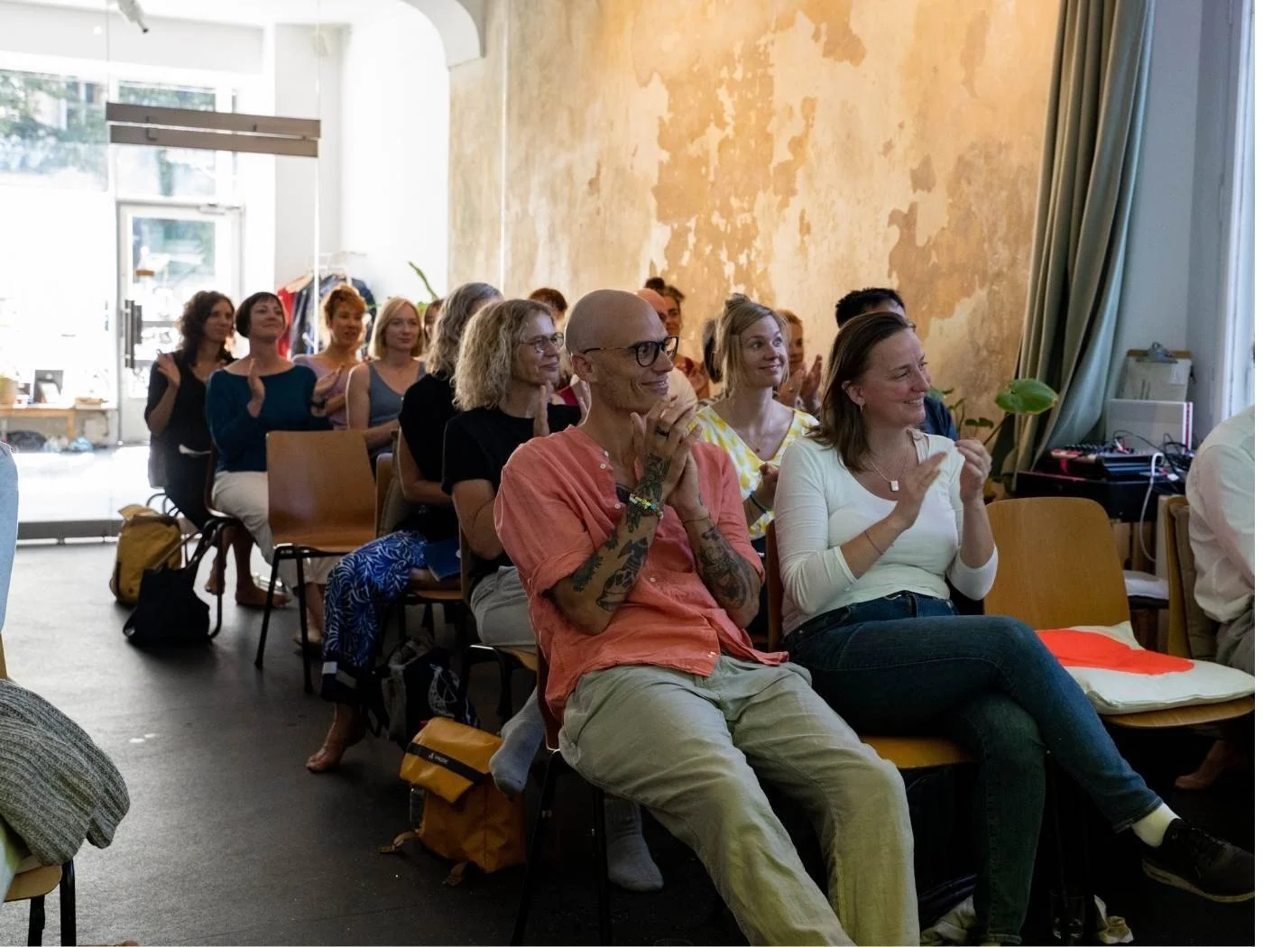 Menschen sitzen in einem Raum und applaudieren bei einer Veranstaltung. Im Vordergrund ein Mann und eine Frau. Im Hintergrund eine Gruppe weiterer applaudierender Personen.