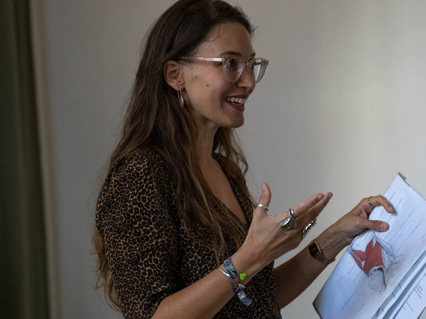 Eine lachende Frau mit langen braunen Haaren und Brille hält ein Skript oder Dokument in der Hand, trägt mehrere Ringe und Armbänder, ist in einem Raum mit neutraler Wandfarbe.