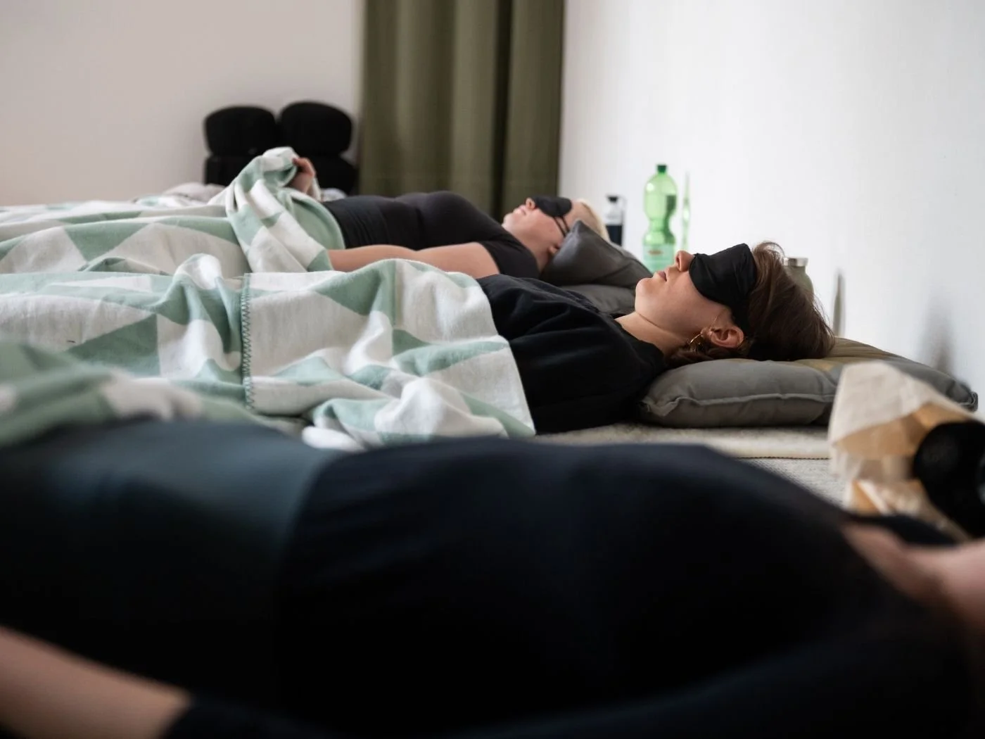 Zwei Frauen liegen schlafend auf Betten, beide tragen Augenmasken, in einem Zimmer mit weißen Wänden und Nachttischen.