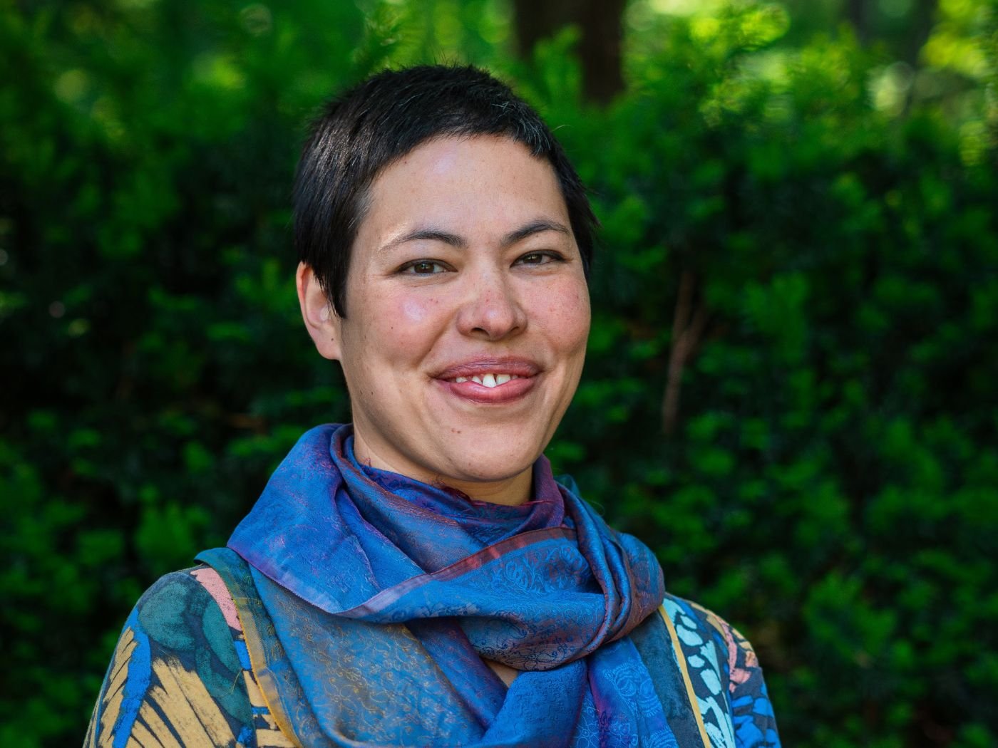 Nahaufnahme einer lächelnden Frau mit kurzen schwarzen Haaren vor einem grünen Wald im Hintergrund.