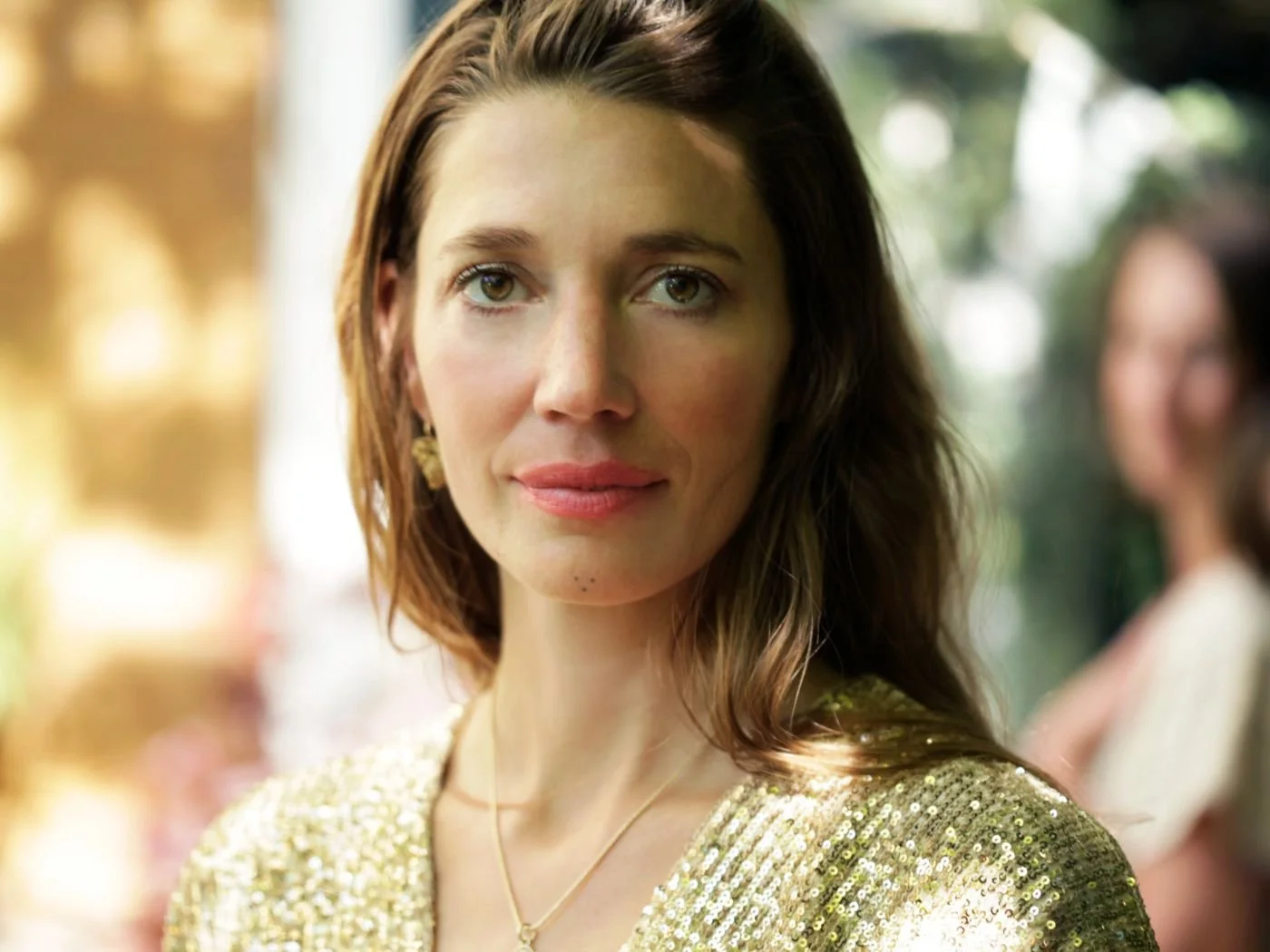 Nahaufnahme einer Frau mit braunen, lockigen Haaren, trägt goldene Kleidung und Ohrringe, im Hintergrund unscharfe Person im weißen Outfit, draußen bei Tageslicht.