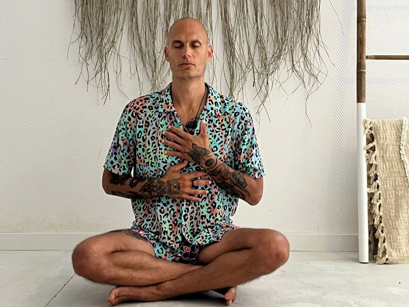 Ein Mann sitzt im Schneidersitz auf dem Boden, hält die Hand auf die Brust und hat die Augen geschlossen bei einer Meditation oder Atemübung. Er trägt ein bunt gemustertes Hemd und Shorts, hat Tattoos an den Armen und Händen. Der Hintergrund ist schlicht mit Dekorationen und einem Kissen.